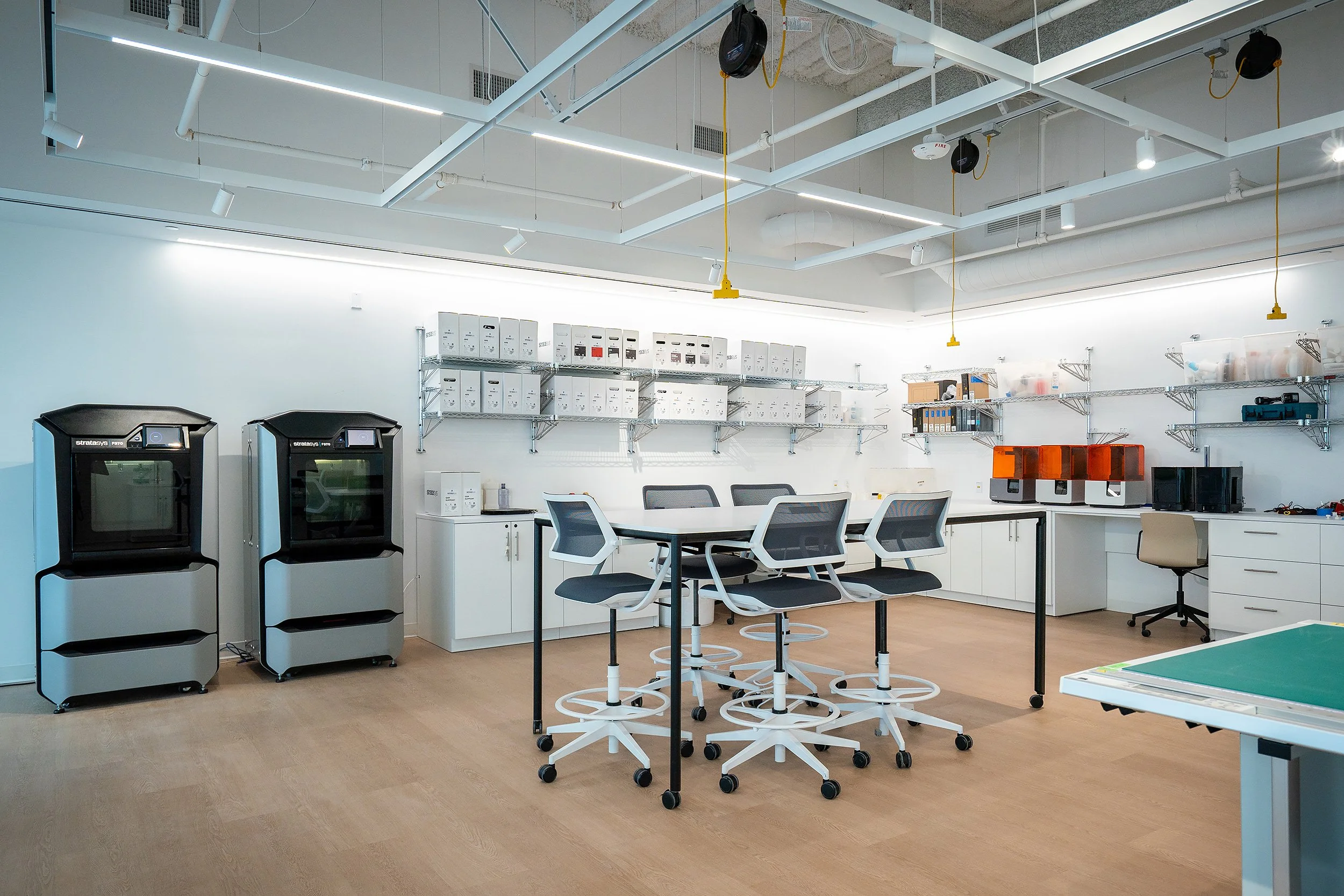 Modern office or lab space with white tables, black and white chairs, shelves with boxes and files, and 3D printers. Ceiling has exposed pipes and lighting.
