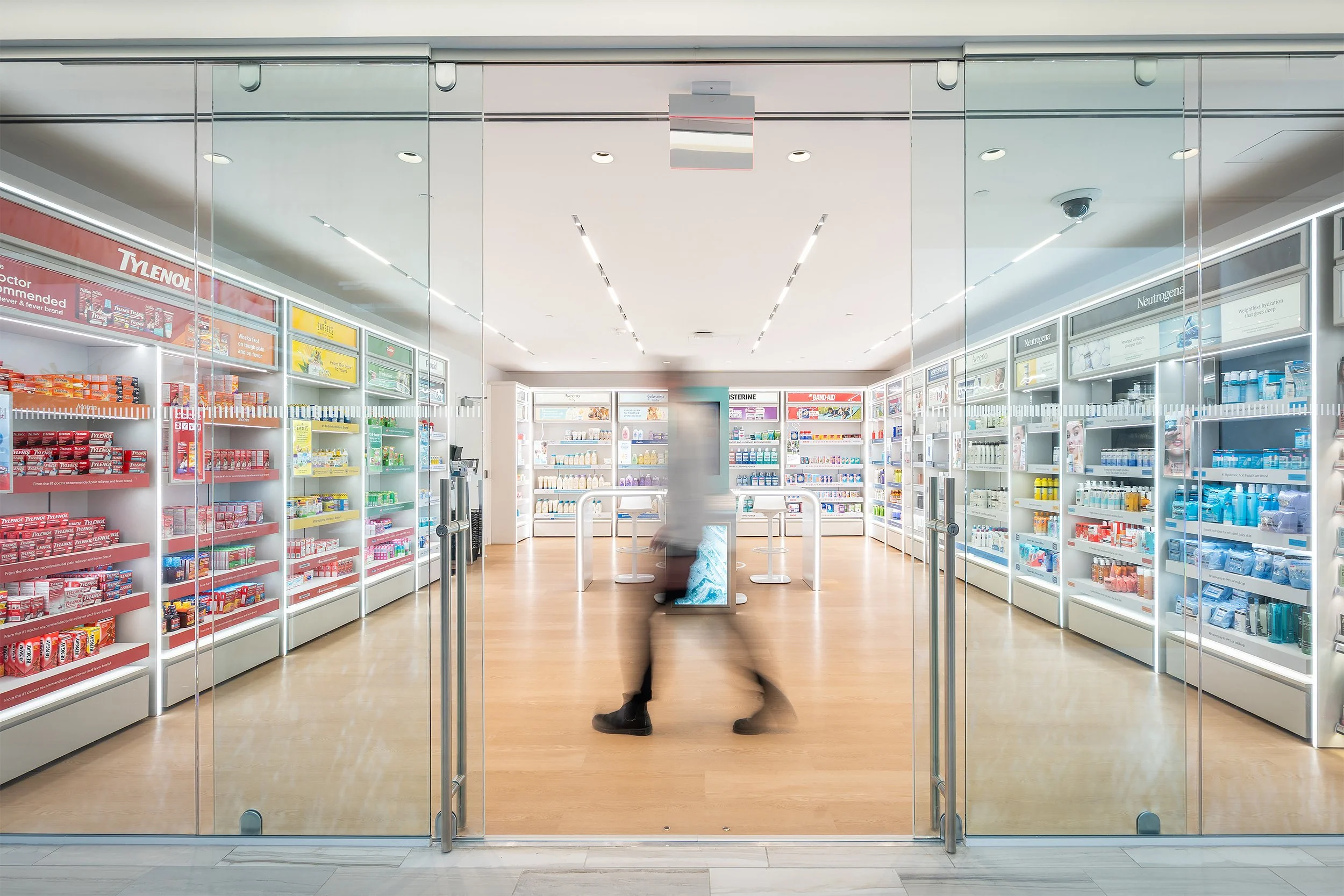 View of a pharmacy or drugstore interior seen through glass doors with shelves of medicines and health products, and a person walking inside.