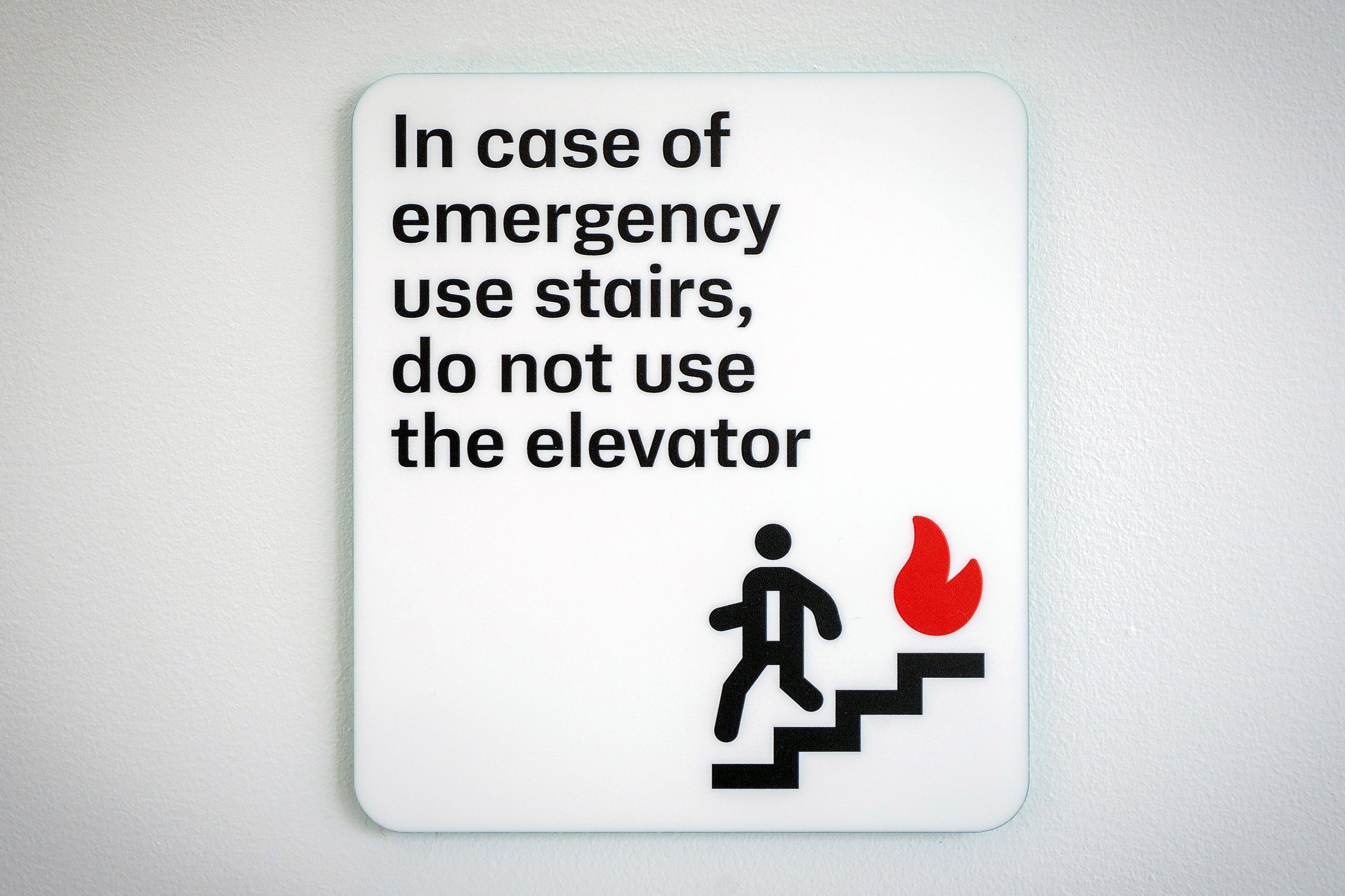 Sign on white wall with instructions for emergency use of stairs and elevator, featuring icons of a person walking down stairs, a fire symbol, and a person at an elevator.