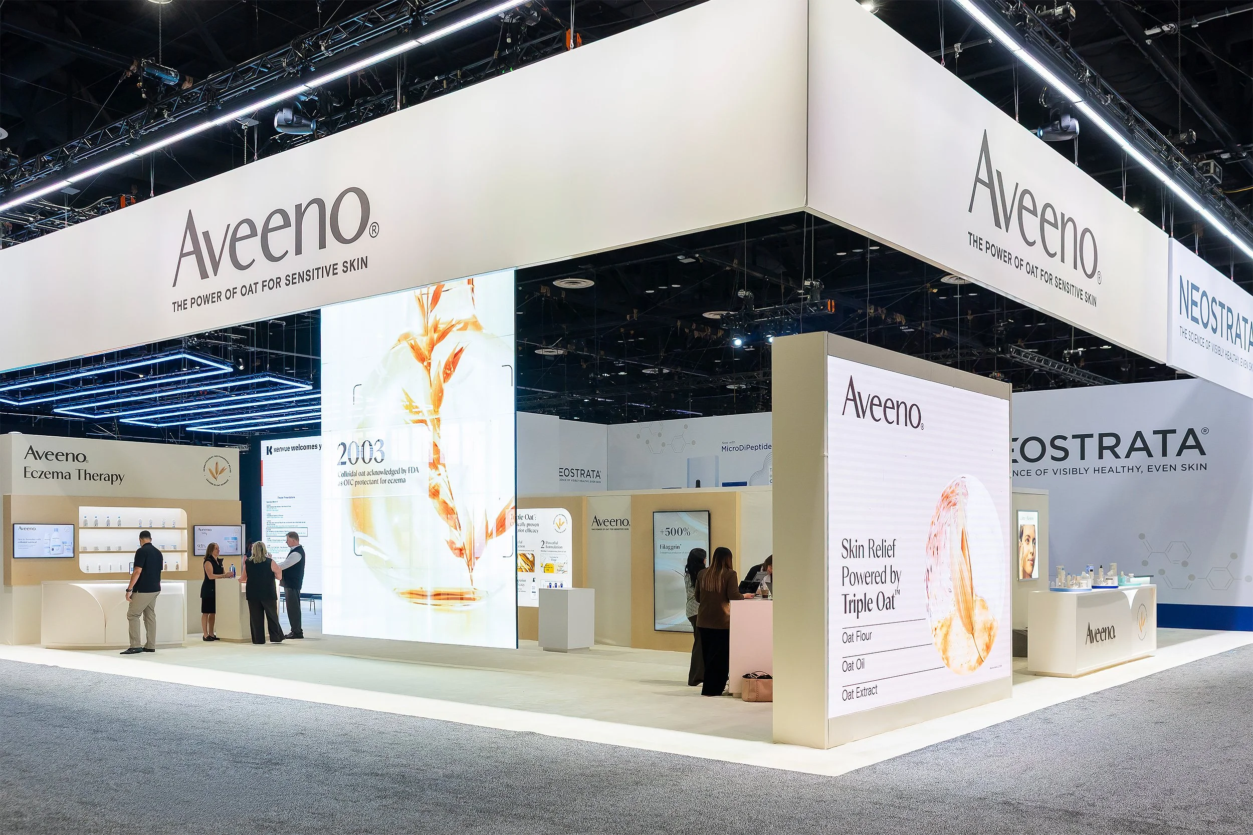 A trade show booth for Aveeno skin care products with large display screens and signage promoting products for sensitive skin, featuring several people browsing and interacting at the booth.
