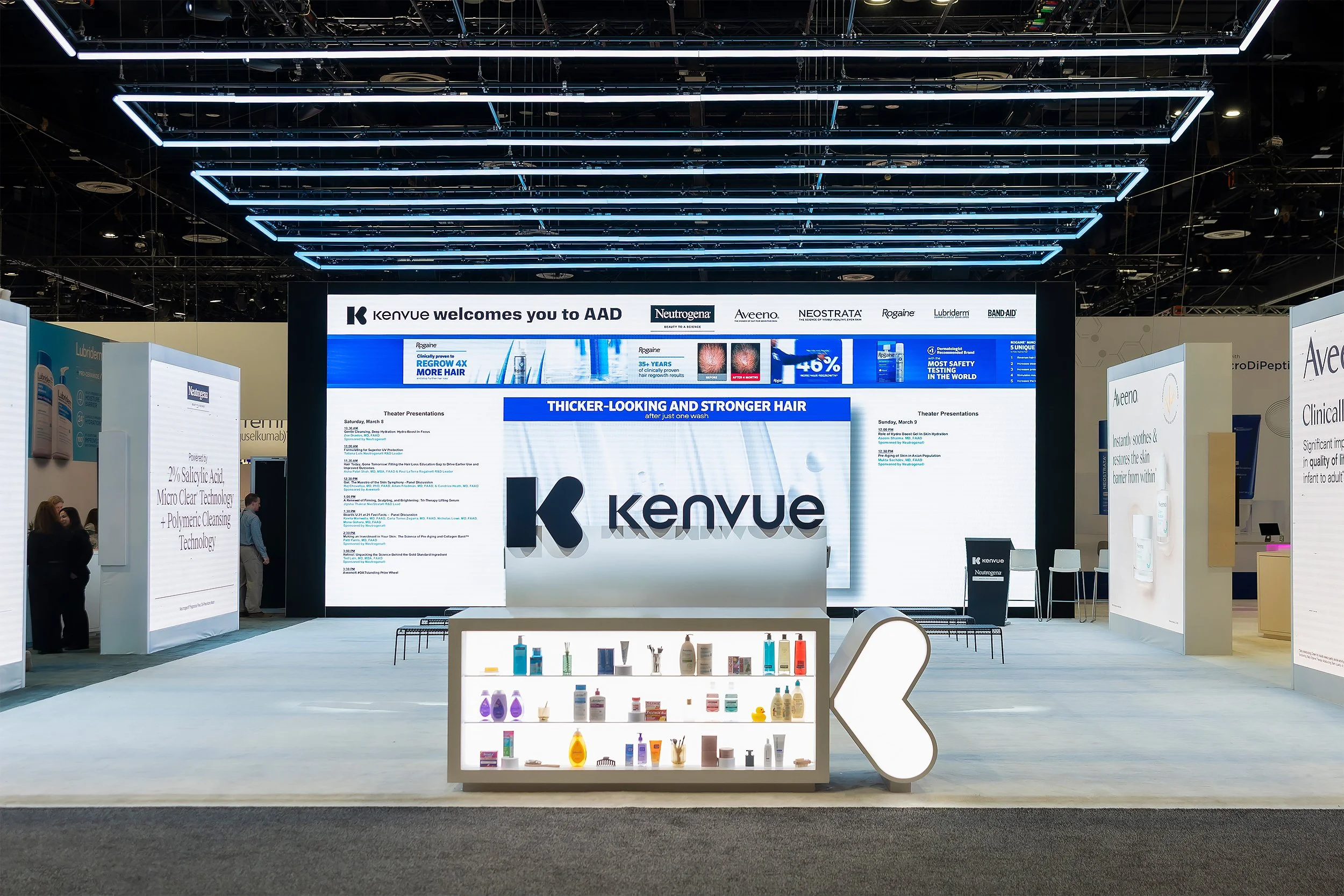 Trade show booth with large digital screen displaying brand names like Kenveu, Neutrogena, Aveeno, and Neostrata. The booth features product displays and promotional signage related to hair and skincare.