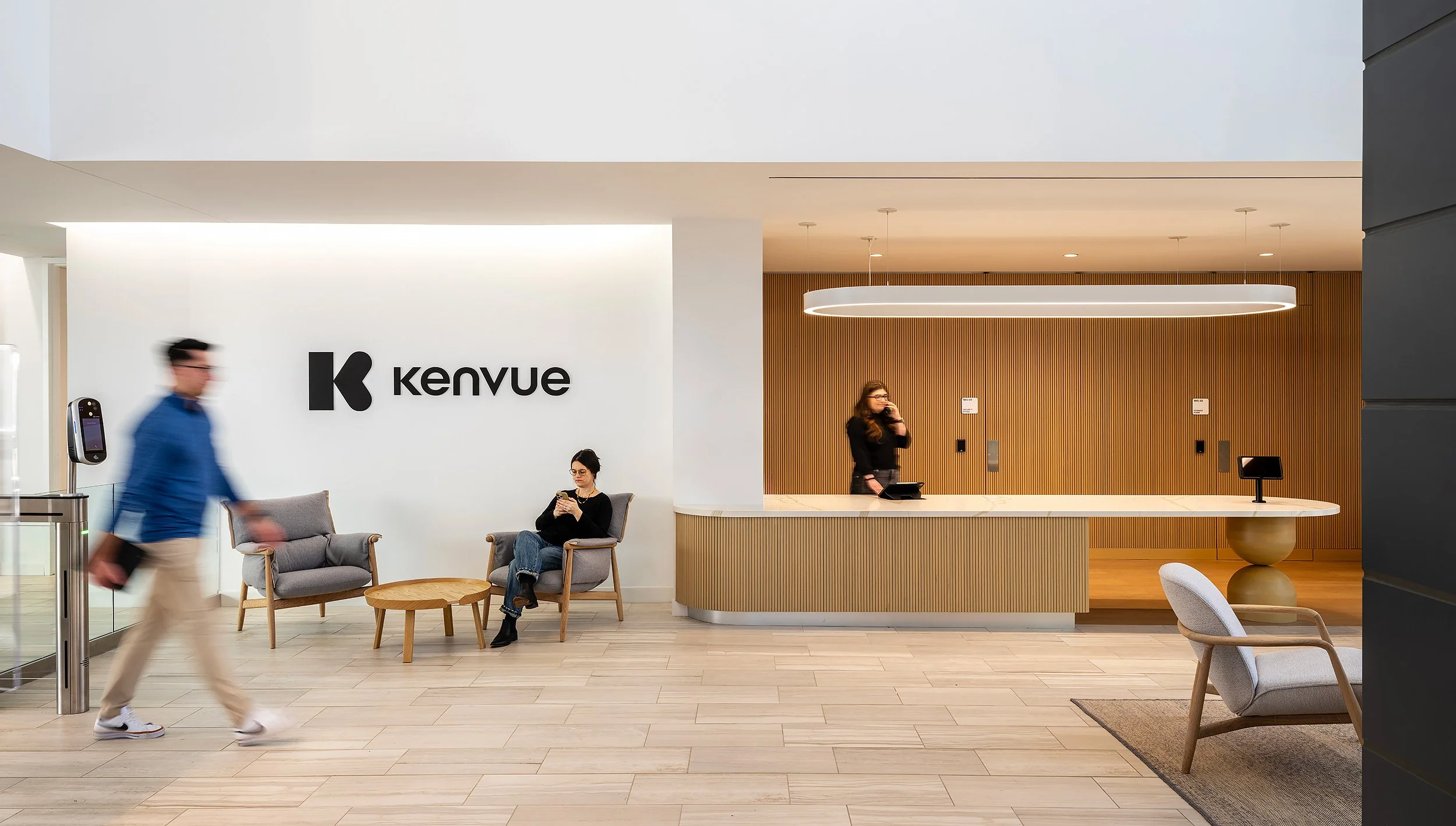 Modern office reception area with a white wall displaying a black logo 'kenvue', wooden accents, a curved front desk, a woman at the desk, a woman in black seated using her phone, an out-of-focus man walking, and chairs in the waiting area.