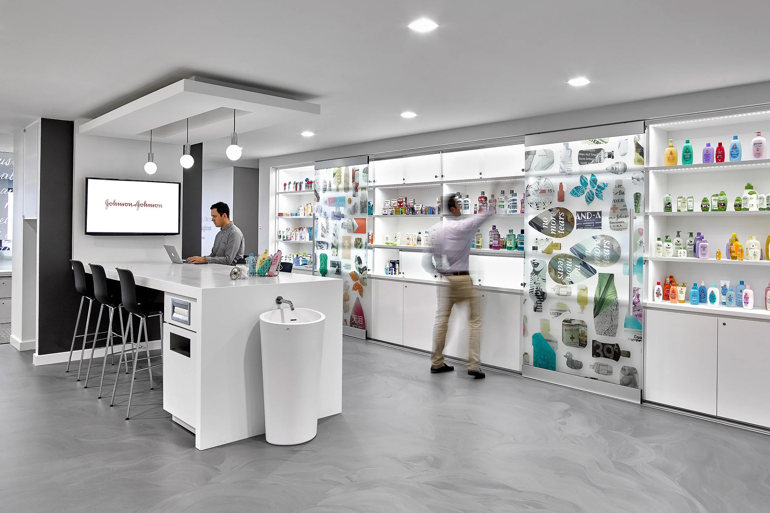 A modern pharmacy interior with shelves stocked with various health and beauty products. A man is reaching for items on a shelf, and another person is working at a counter with a man working on a laptop.