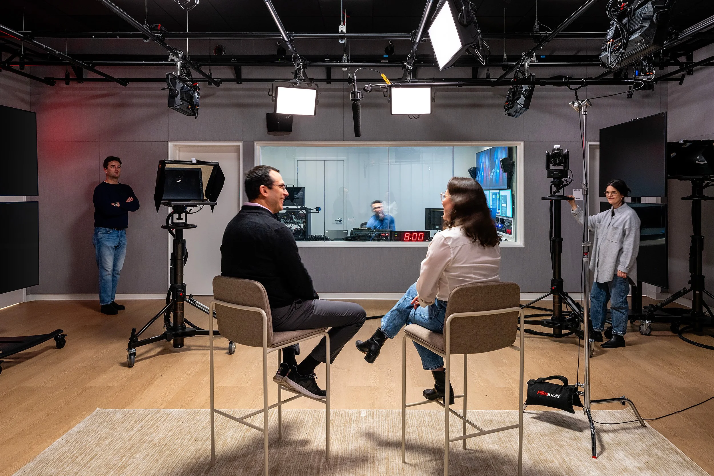 Two people sit and face each other in a studio surrounded by filming equipment, with a large window behind them revealing a control room. Two others stand on either side of the scene, one with arms crossed and the other holding a camera. The setup suggests a filming or interview session.