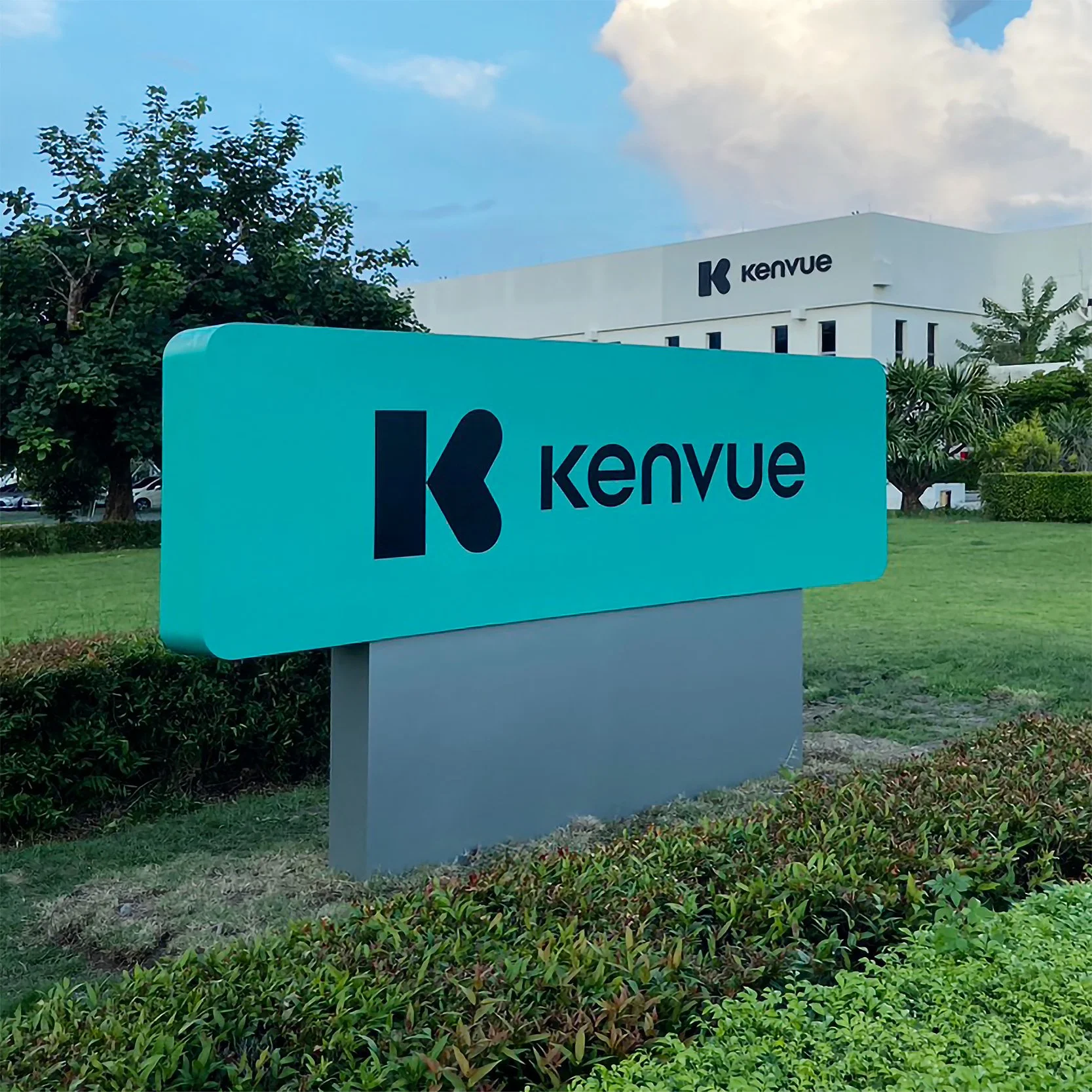 Photo of a building with the Kenvue logo, and a blue sign with the same logo in front of it, surrounded by greenery and trees.