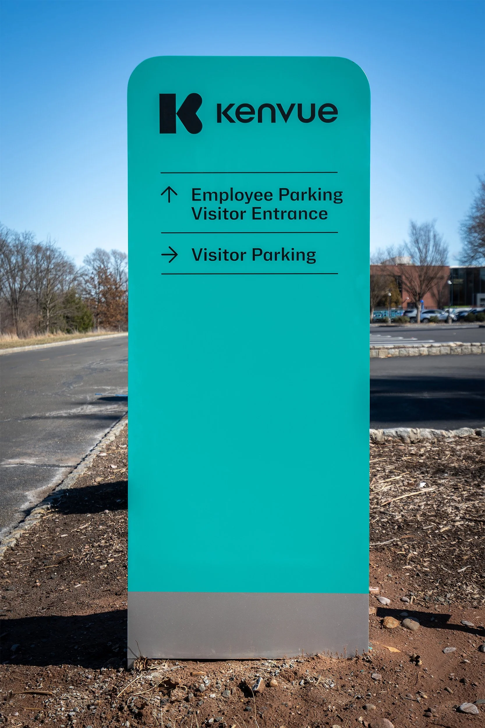 Sign for Kenvue with directions for employee parking and visitor parking, located outdoors on a paved lot with trees and a building in the background.