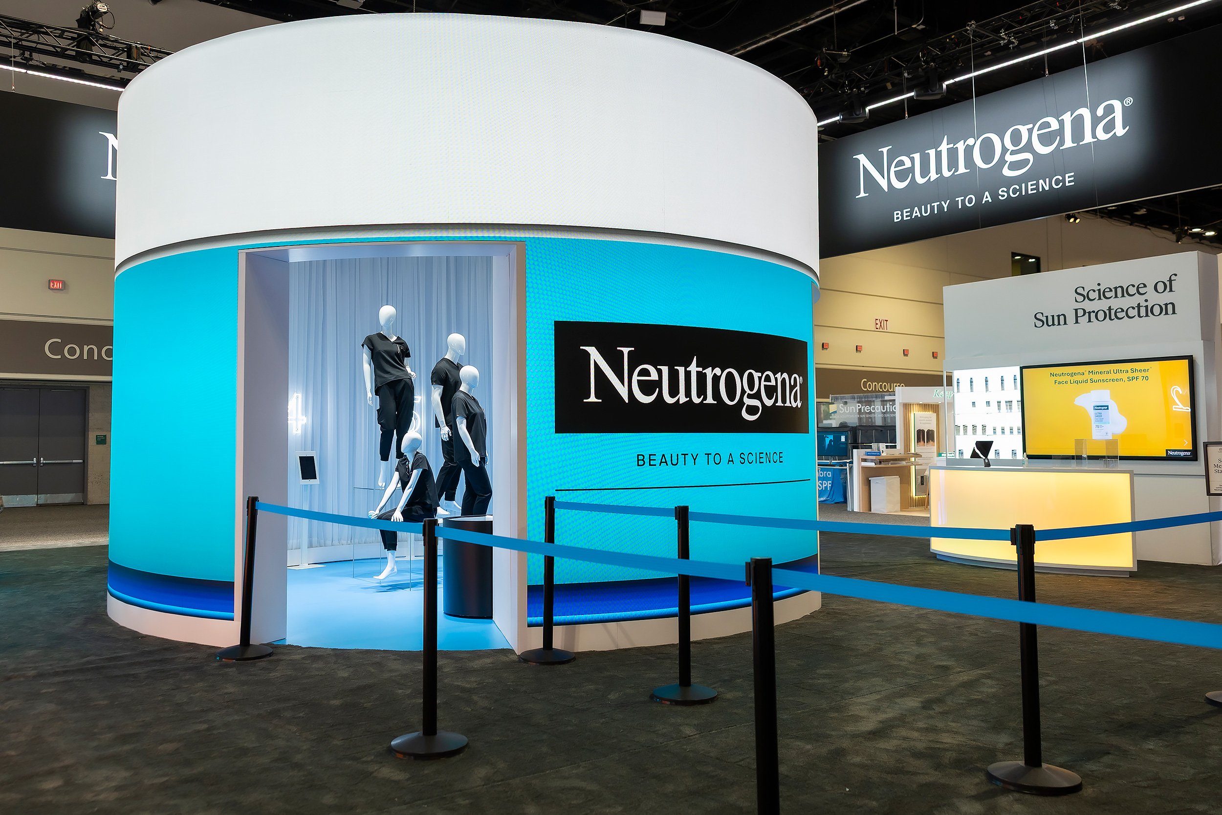 Exhibition booth for Neutrogena with mannequins wearing black outfits, digital screens, and signage displaying the Neutrogena logo and products at a trade show.