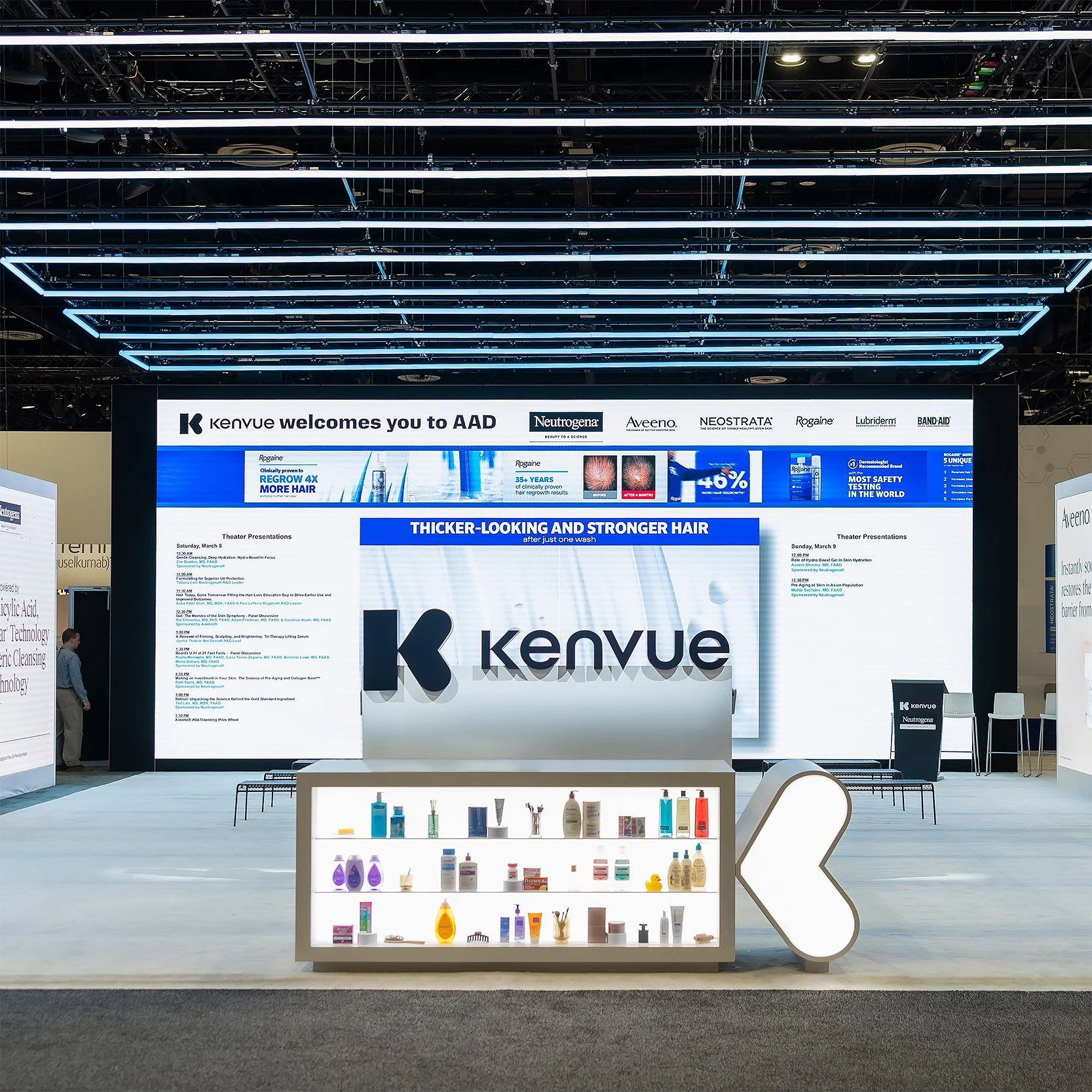 A large display booth at a trade show for Kenveu, featuring an illuminated sign with the brand's logo and informational text about hair care products, including a shelf with various bottles and containers of personal care items.