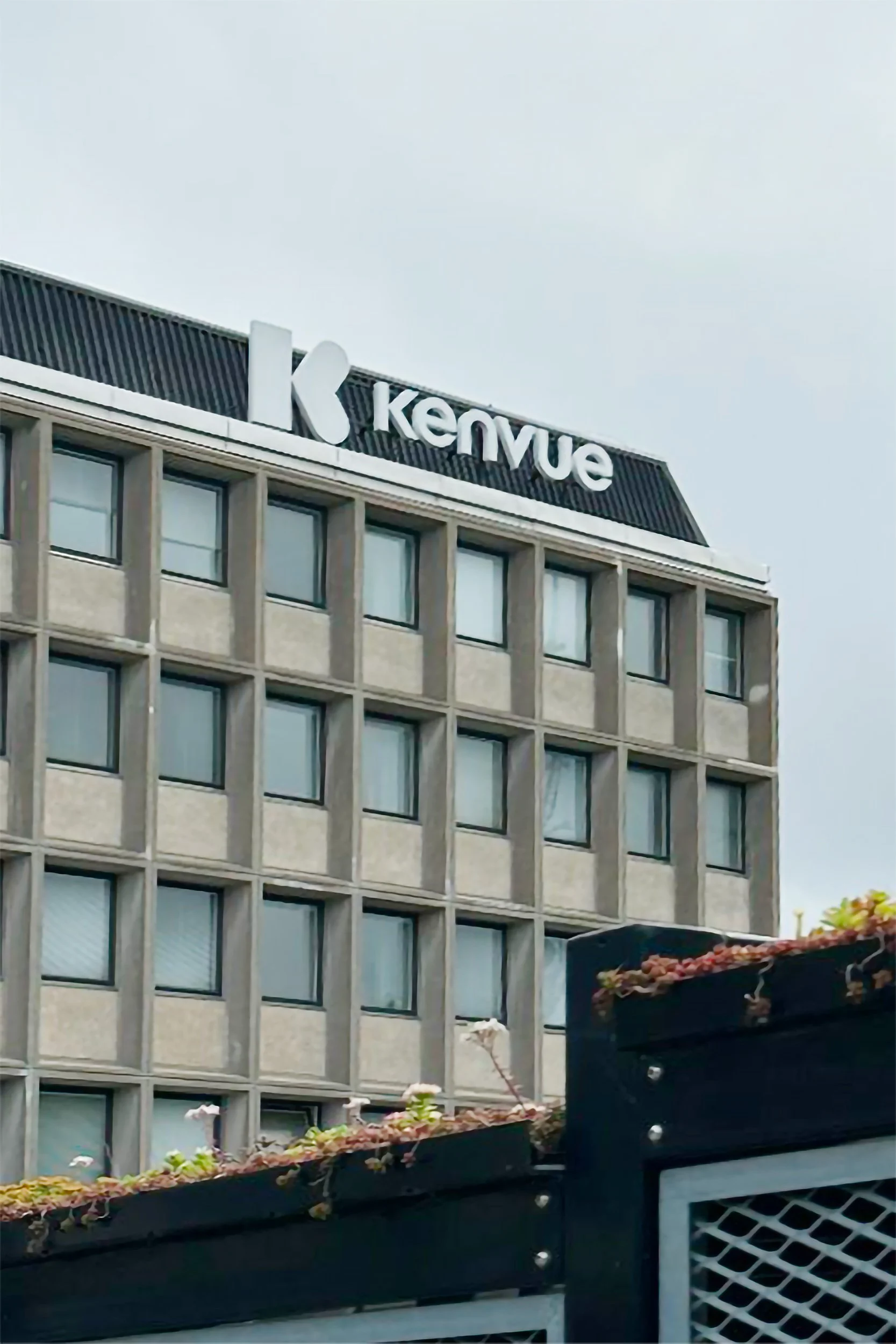 The image shows the top part of a building with the sign 'Kemvue' on the roof, under an overcast sky. The building has a grid of rectangular windows and part of a black rooftop garden with plants.