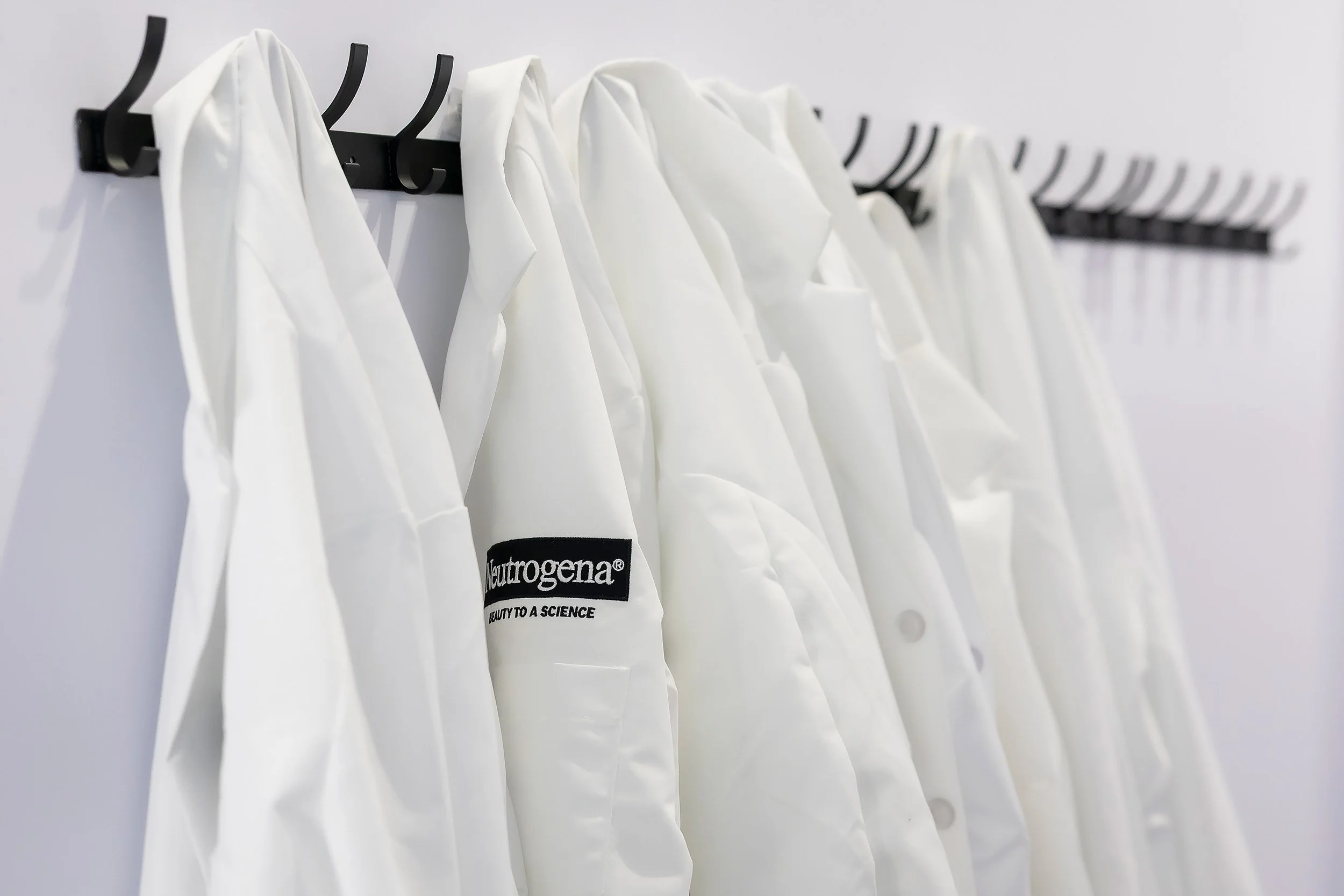 White lab coats hanging on black hooks on a white wall, with one coat displaying a label that reads "Neutrogena" and the slogan "Beauty to a Science."