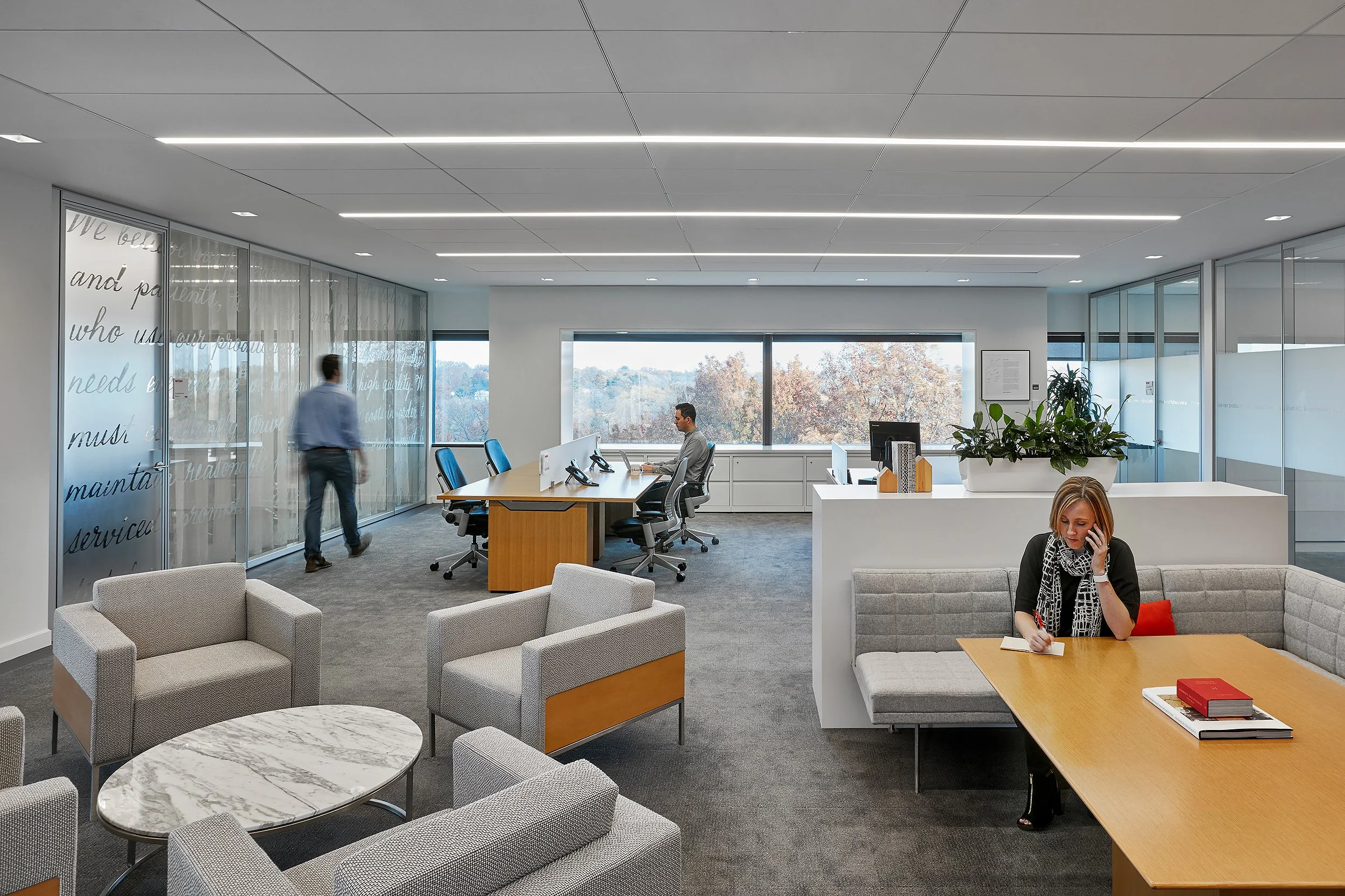 A modern, open-plan office with large windows showing trees outside. The office has a mix of seating including armchairs and a conference table. There are three people: one seated at a desk using a computer, one walking near glass walls, and a woman sitting on a couch talking on her phone.