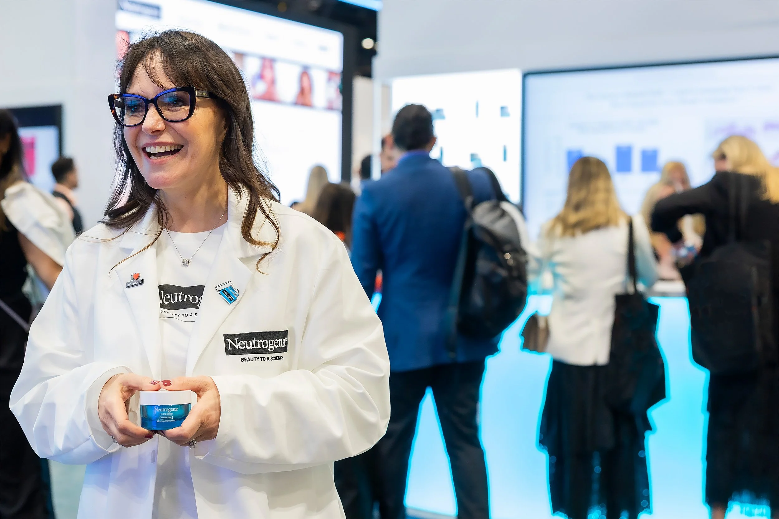 A woman wearing glasses and a white lab coat with Neutrogena branding smiling and holding a container of Neutrogena product. She is at a busy event or conference, with other people in the background and digital screens displaying information.
