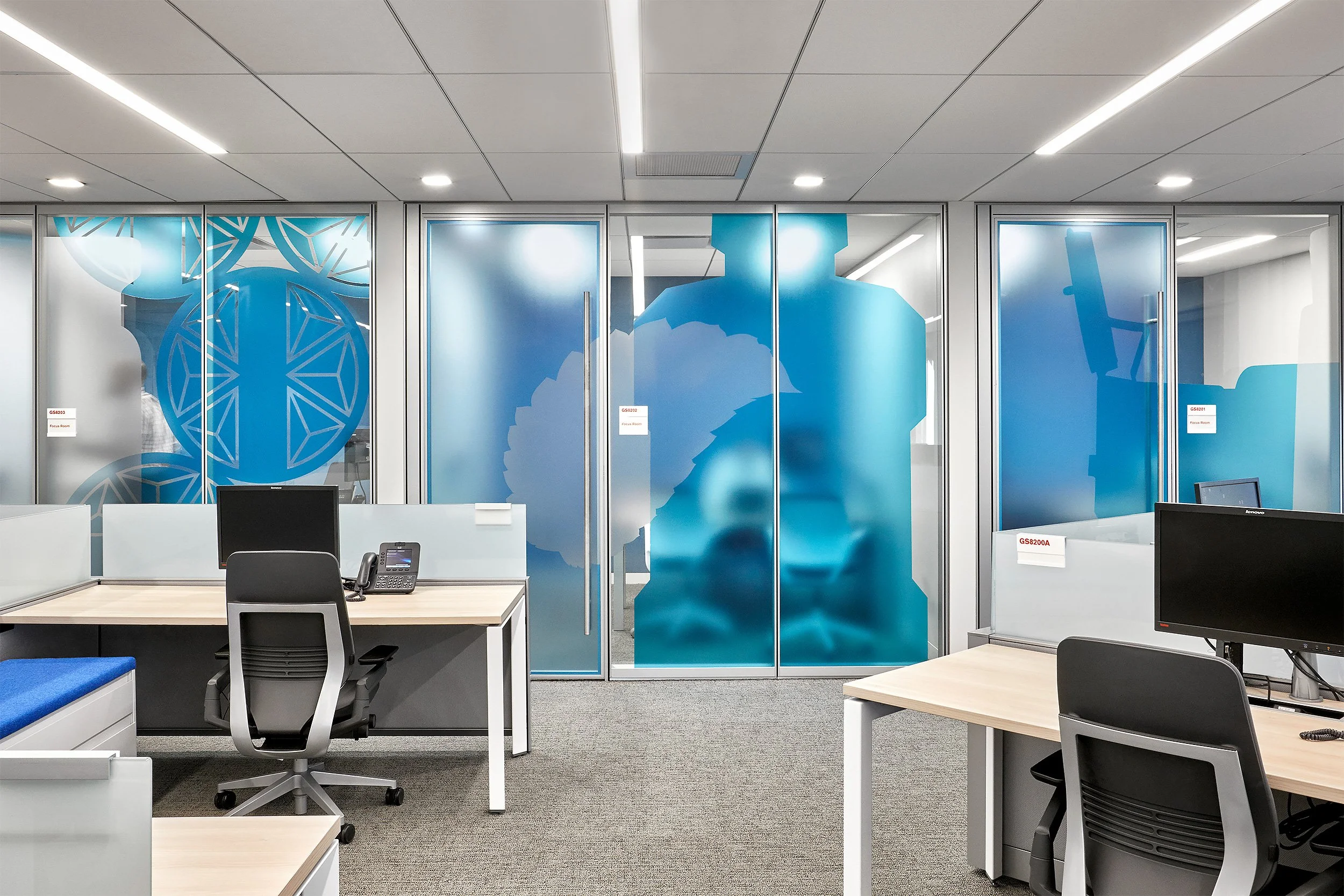 Modern office space with desks, office chairs, computer monitors, and glass doors with blue graphics of scientific symbols and molecules.
