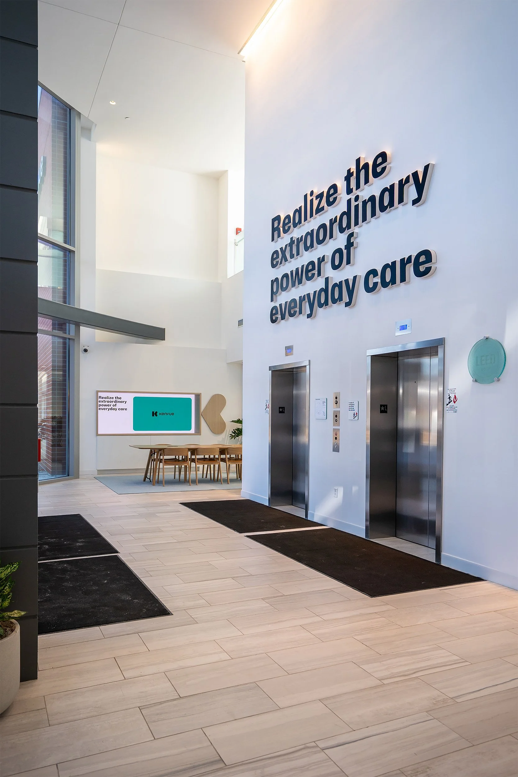 Modern indoor space featuring two elevators, a seating area with a round table and chairs, and a large wall with the words 'Realize the extraordinary power of everyday care' in bold letters. The area has large windows, light-colored wood flooring, and black welcome mats.