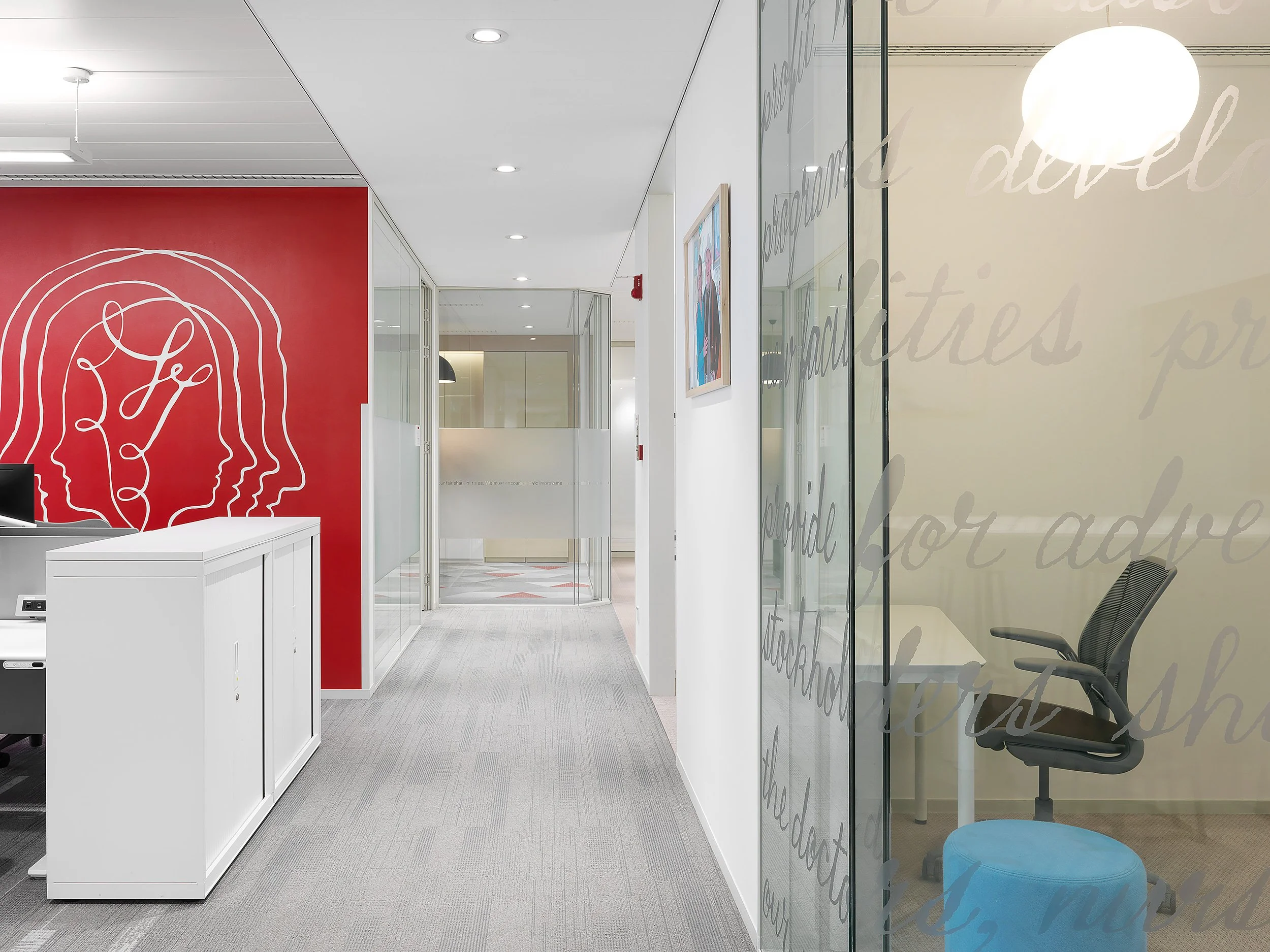 Modern office hallway with glass walls, seating area, and artwork, featuring a red accent wall with a line drawing of a head with interconnected lines.