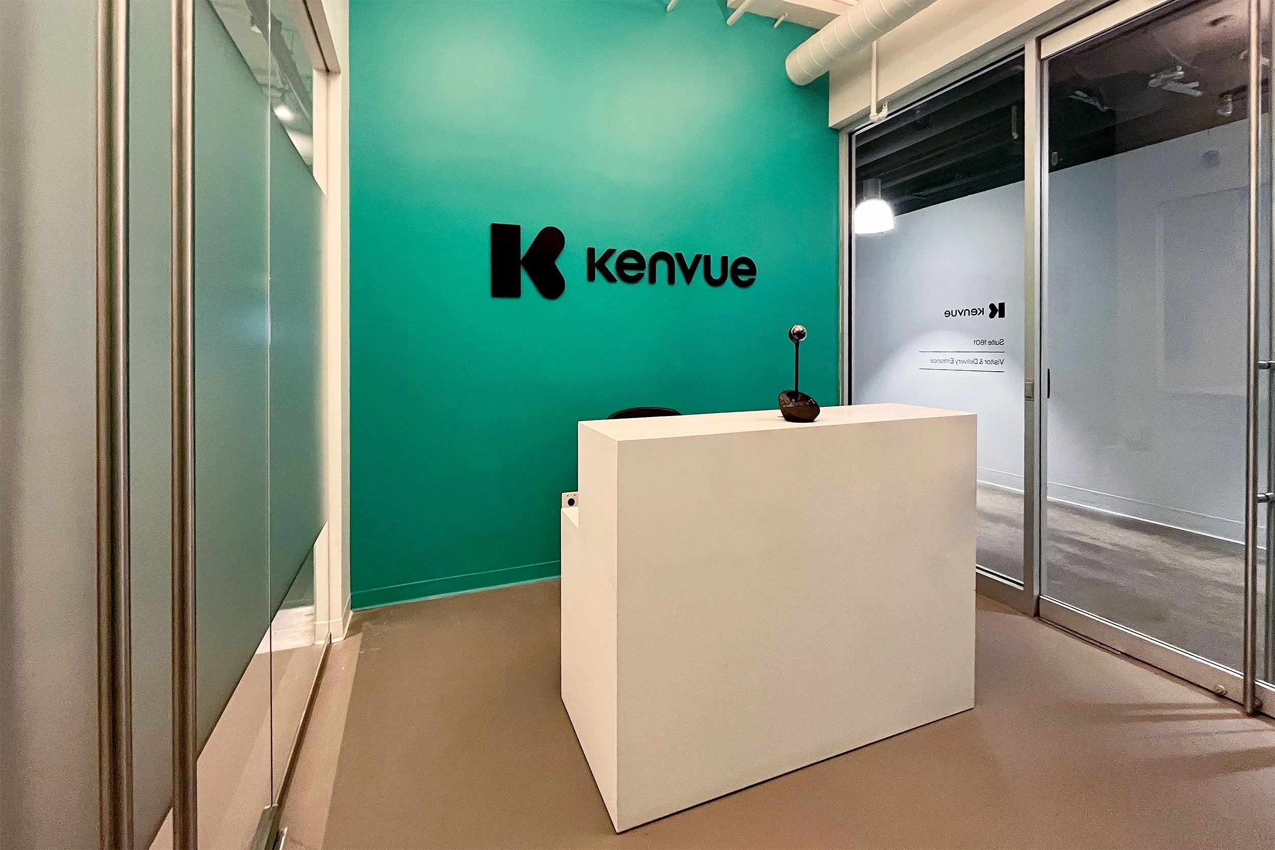 Reception desk in an office with a teal accent wall displaying the Kenvue logo, glass wall panels, and a microphone on the desk.