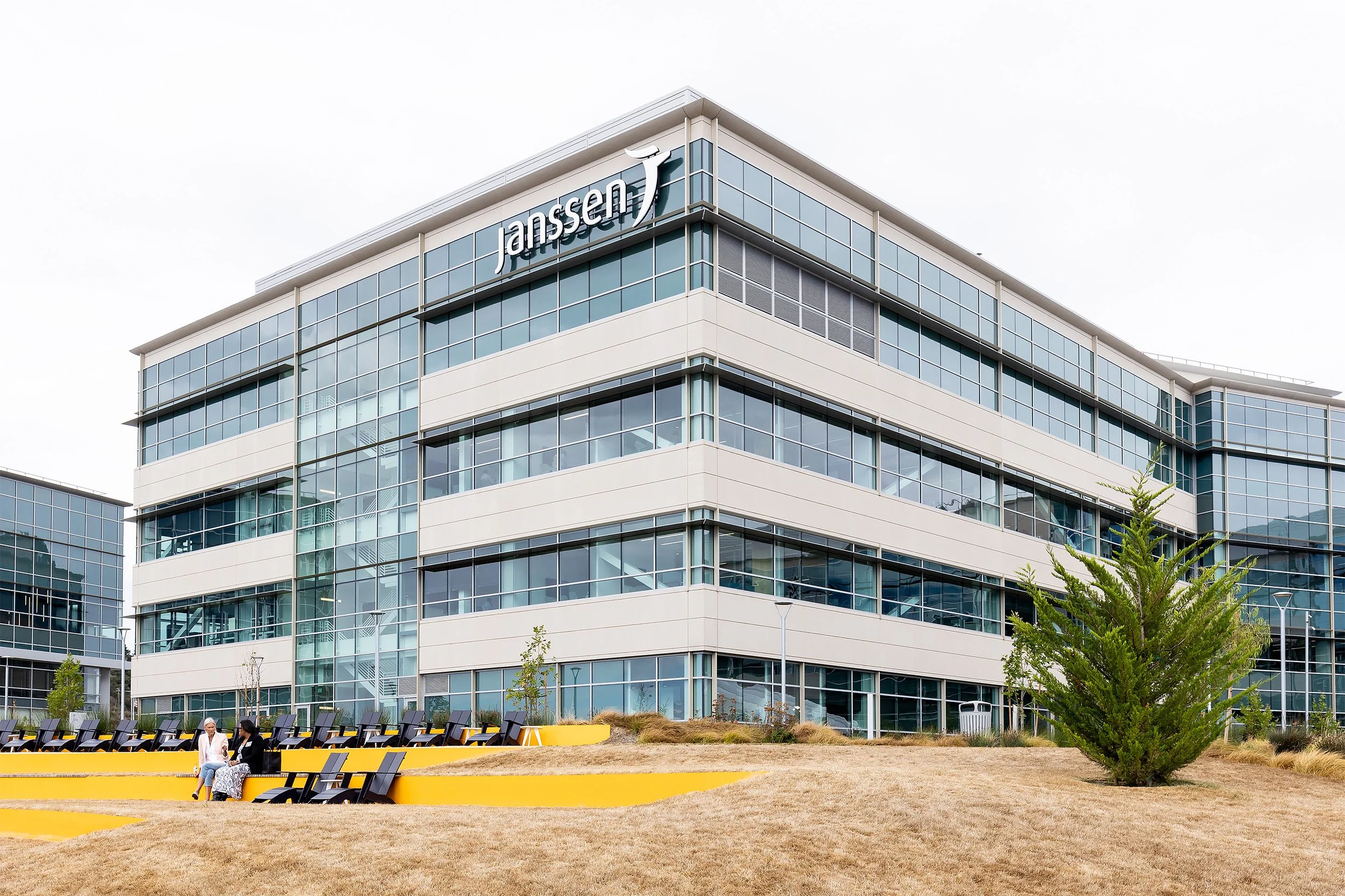 A modern multi-story office building with glass windows and the logo 'Janssen' on the upper corner of the building. There is an outdoor seating area with yellow and black benches and two people sitting and talking. Small trees and landscaped grass are in the foreground.
