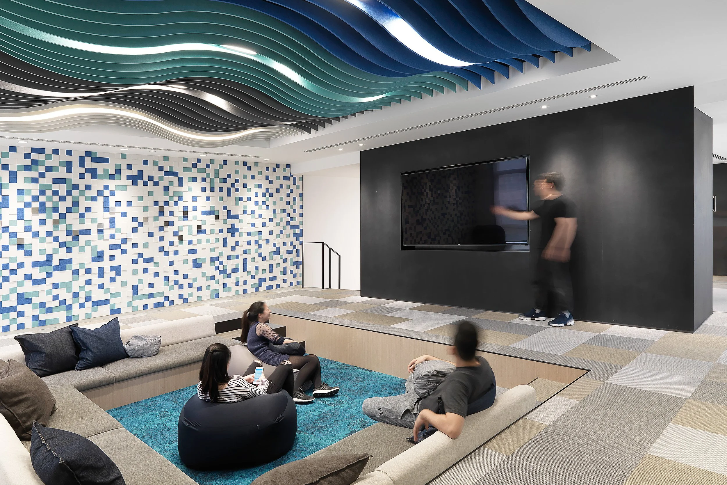 A modern indoor lounge with a ceiling featuring wavy, layered panels in shades of blue, green, black, and white. The wall on the left has a mosaic pattern of blue and white tiles. There is a black wall-mounted television and a person standing beside it. Several people are sitting and lying on beige sofas and bean bags, some using their phones.
