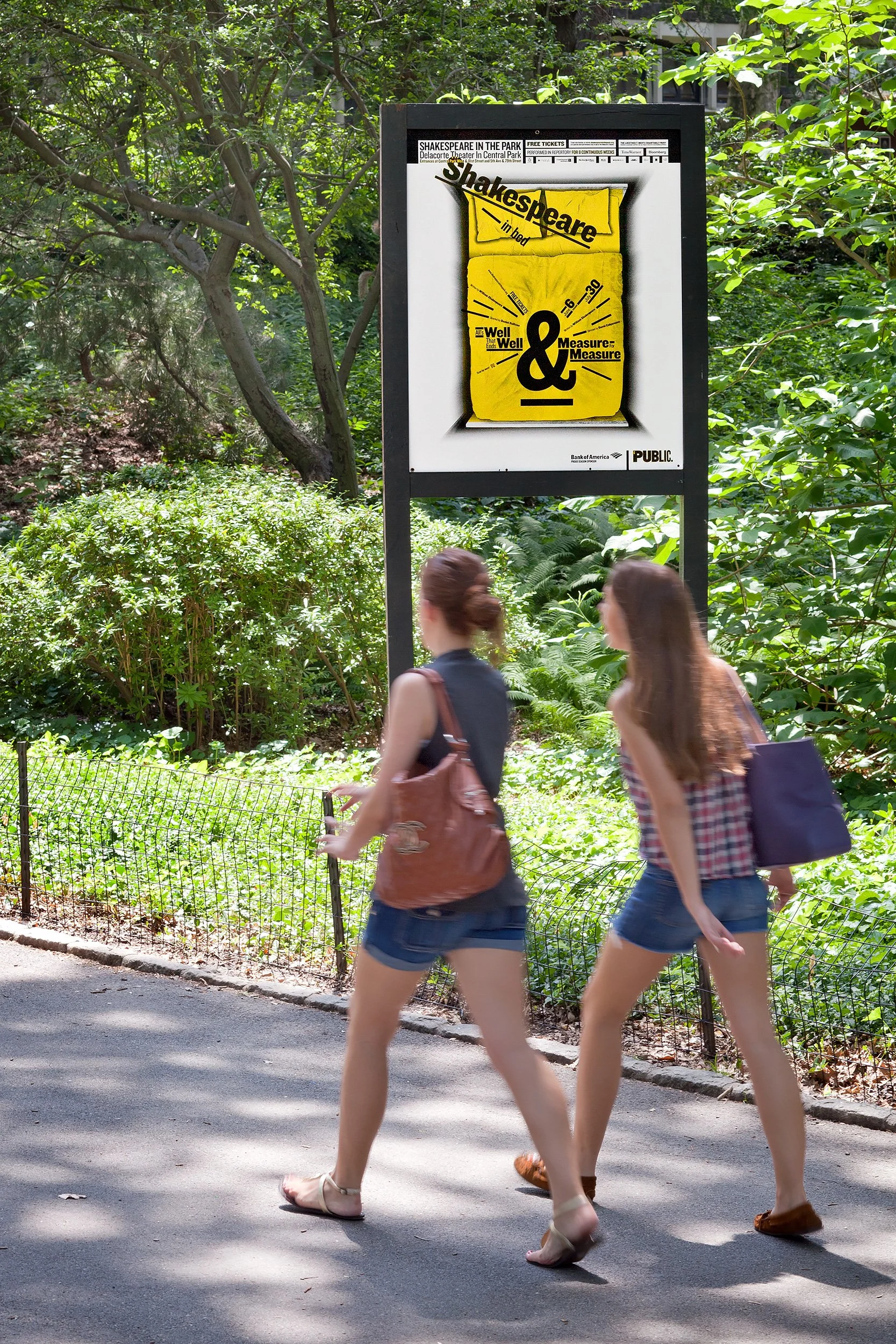 Two women walk along a sidewalk in front of a lush green park area, wearing casual summer clothing.