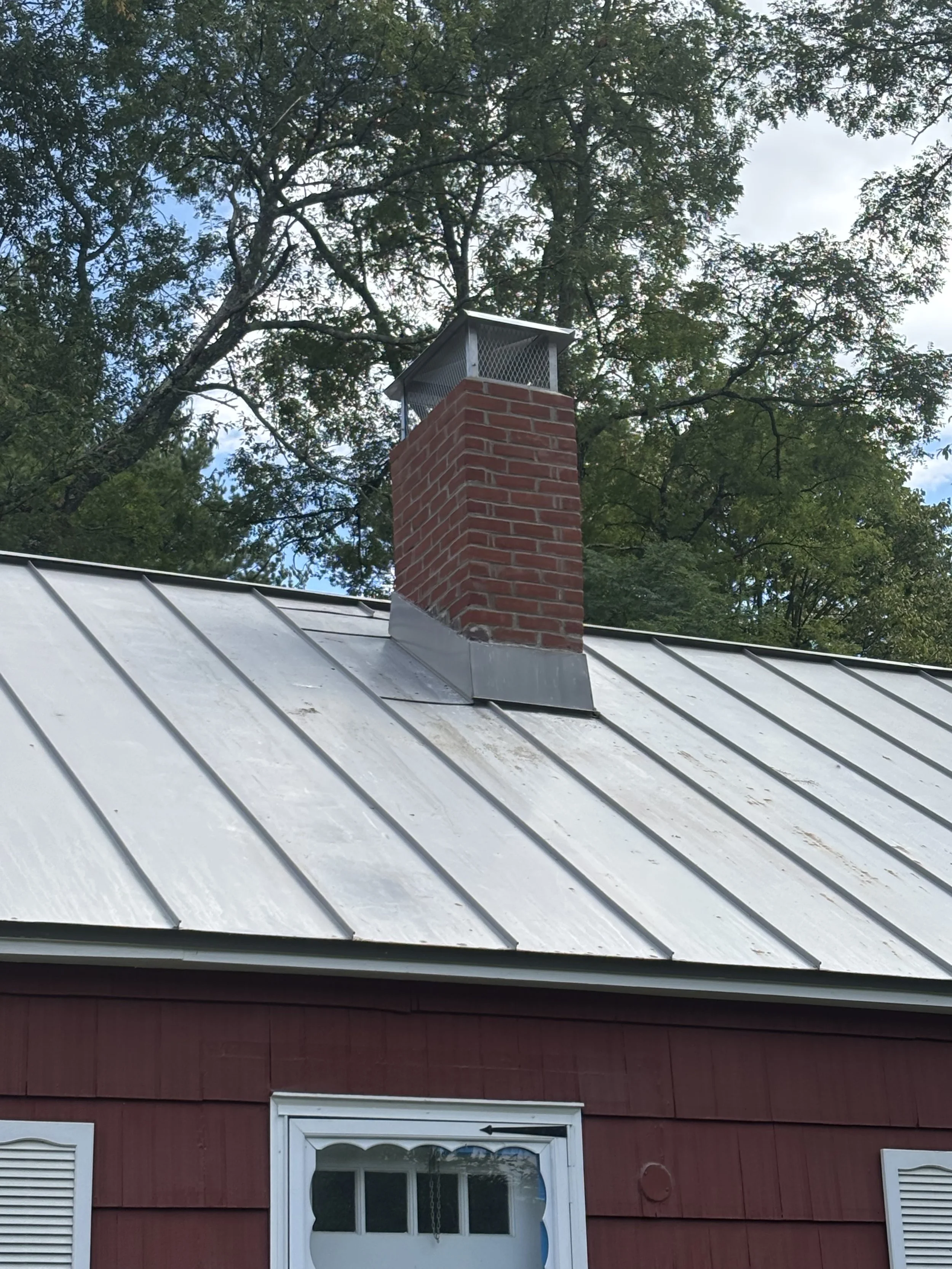 Metal roof with vent pipe and brick chimney on a red house with two windows, surrounded by trees and a partly cloudy sky.