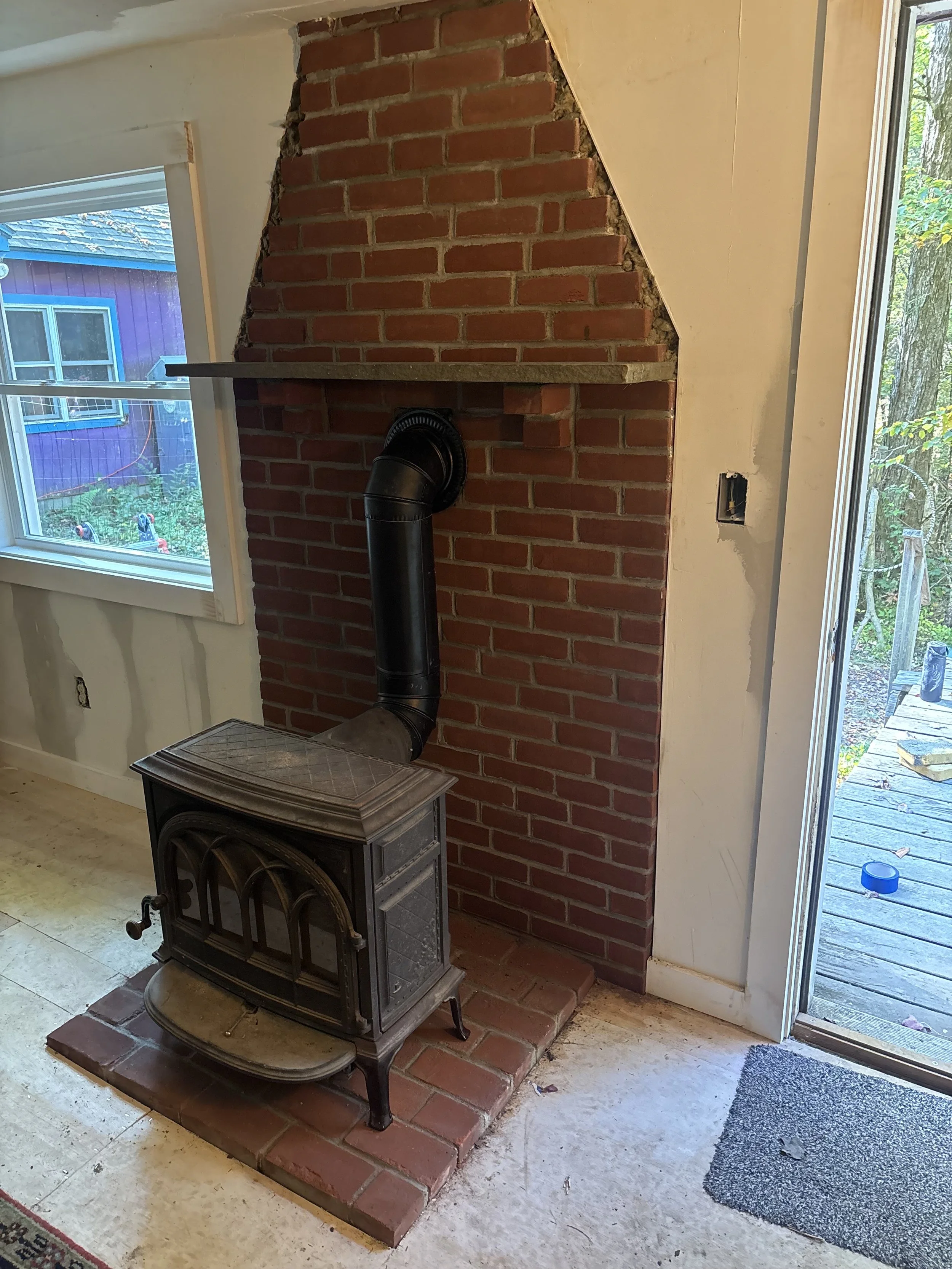 A wood-burning stove in front of a brick chimney with a metal shelf on top, inside a room with a window and a sliding door leading outside to a deck.