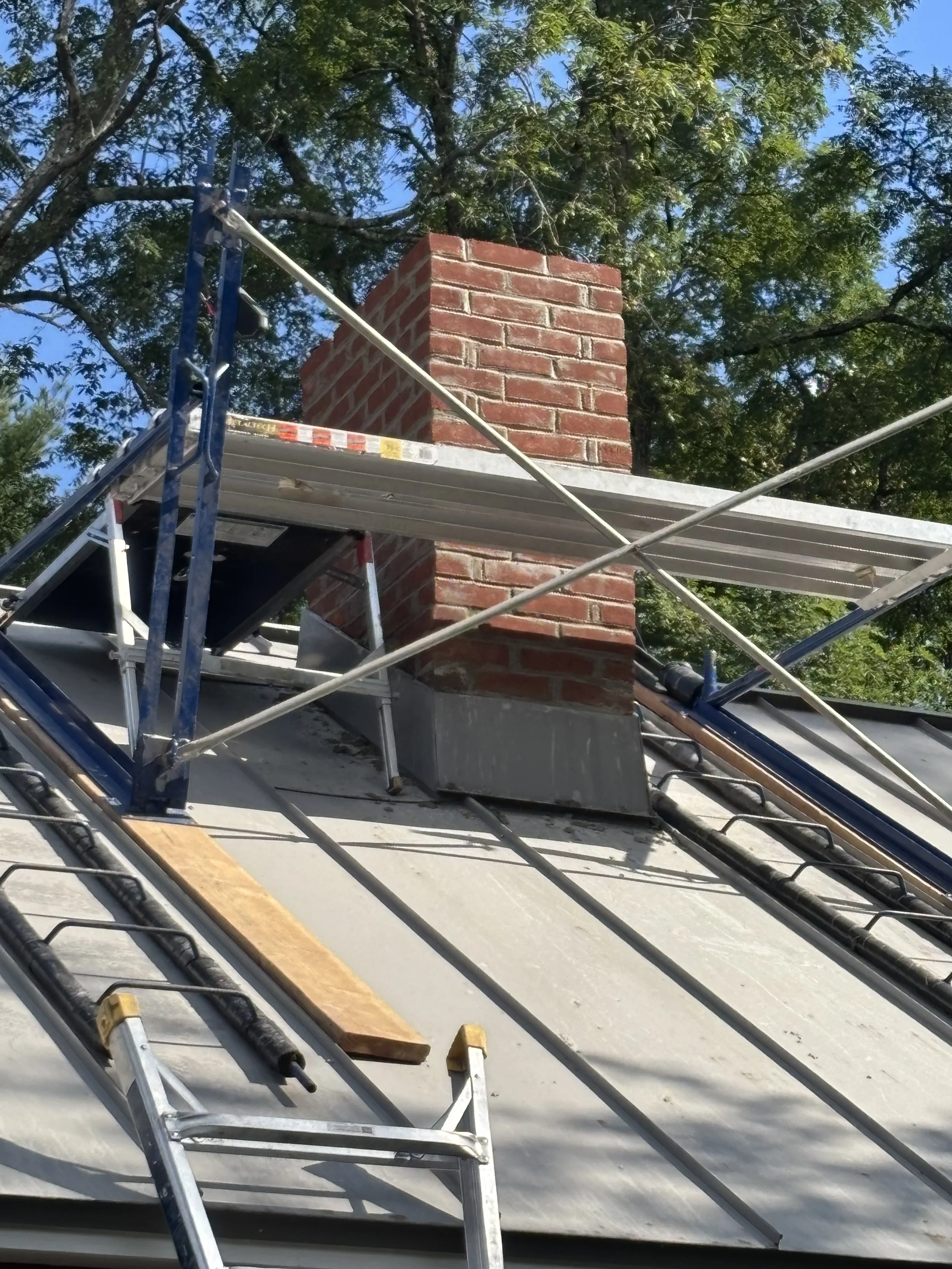 View of a chimney with a red brick exterior on a roof, surrounded by scaffolding and construction tools, with a background of green tree foliage and a partly cloudy sky.
