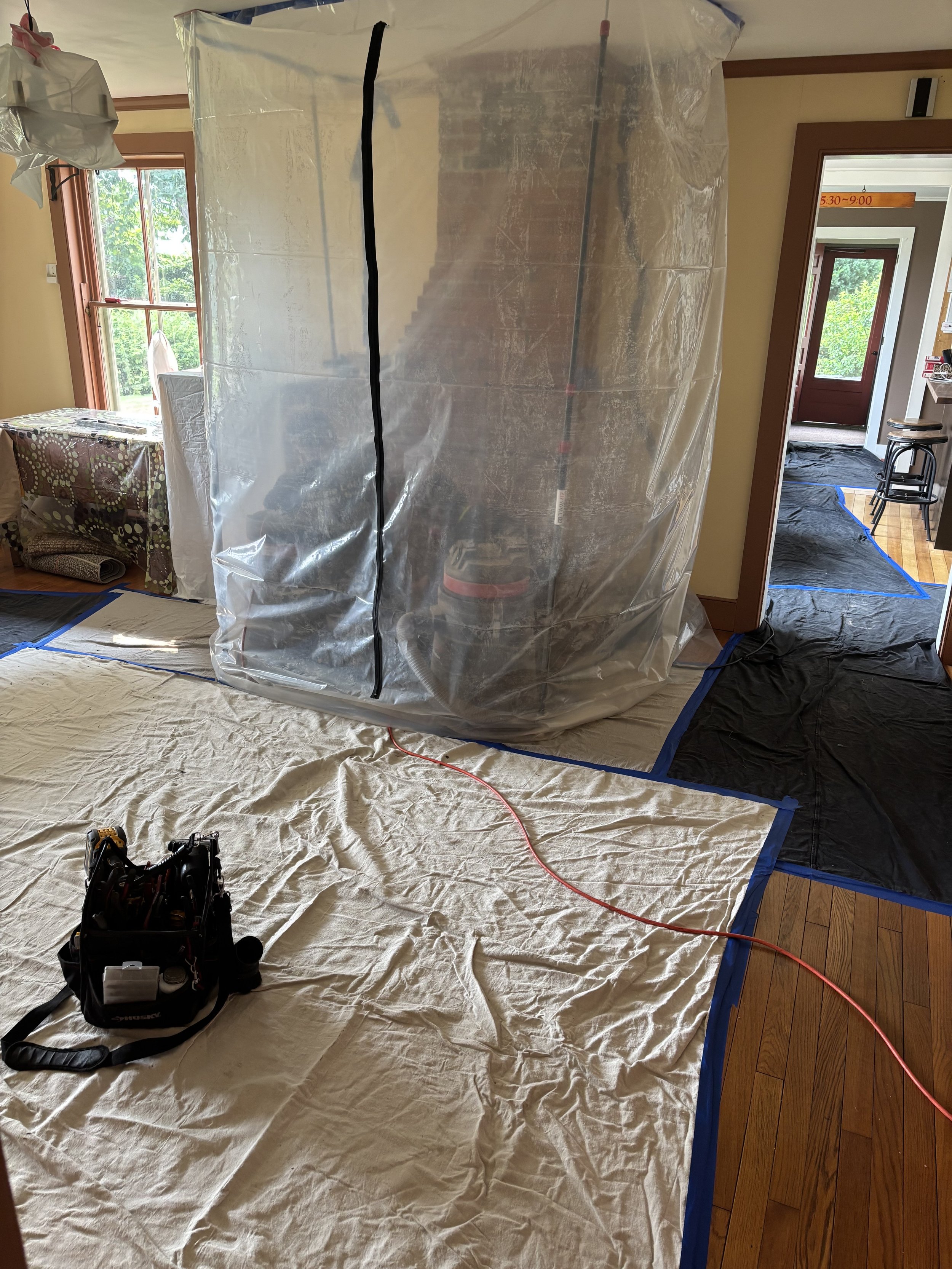 Room mid way through chimney cleaning service with drop cloth covering the floor and plastic sheeting enclosing the fireplace area, masks on the floor, and construction equipment.