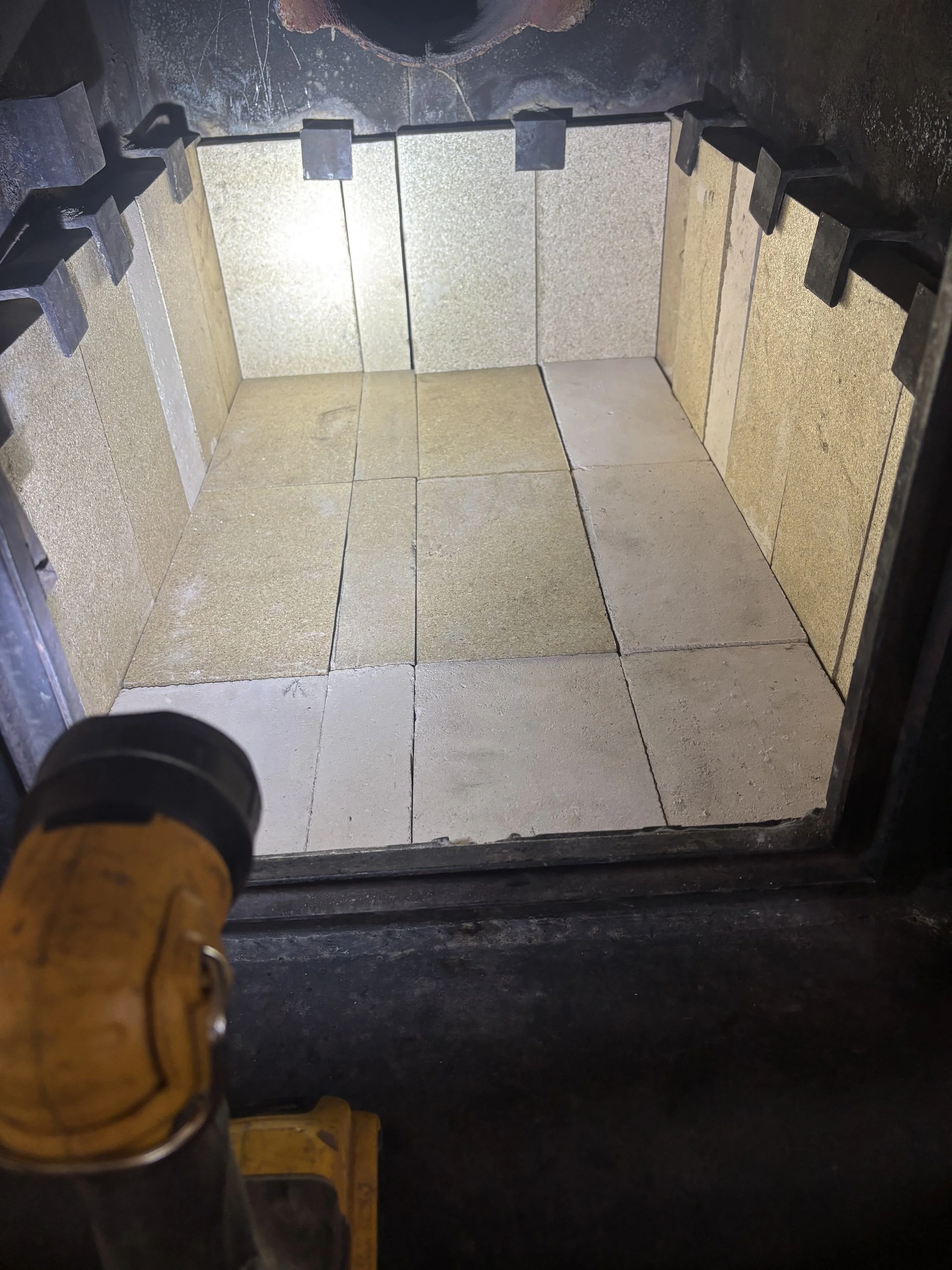 Interior of a freshly cleaned fireplace with beige and light yellow tiles arranged on the floor and walls, seen from the opening with a torch pointed inside.