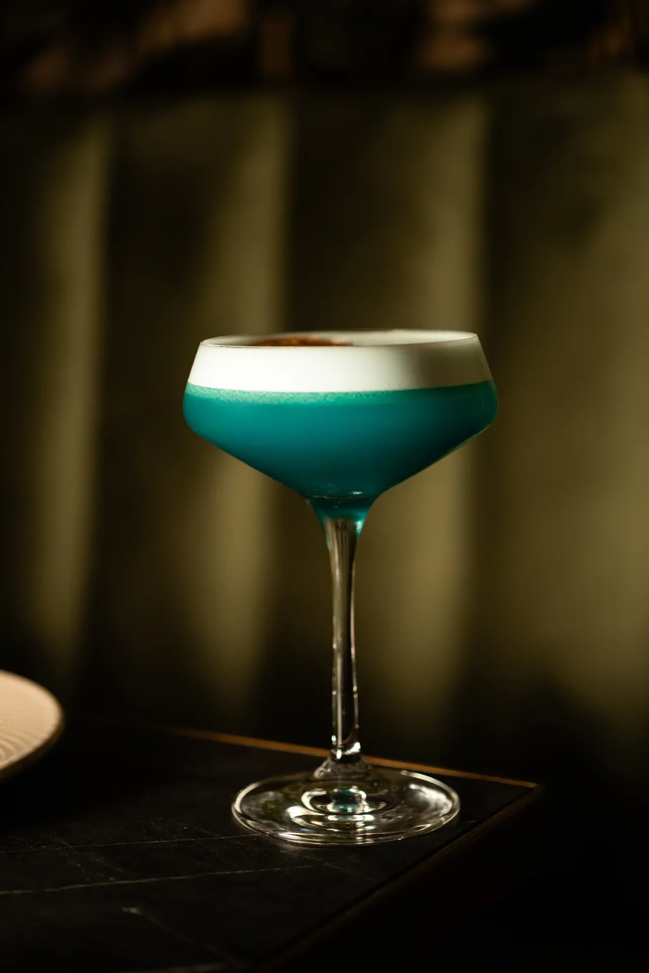 A cocktail glass with a layered blue and white drink, topped with a layer of foam, placed on a dark surface with a blurred background.