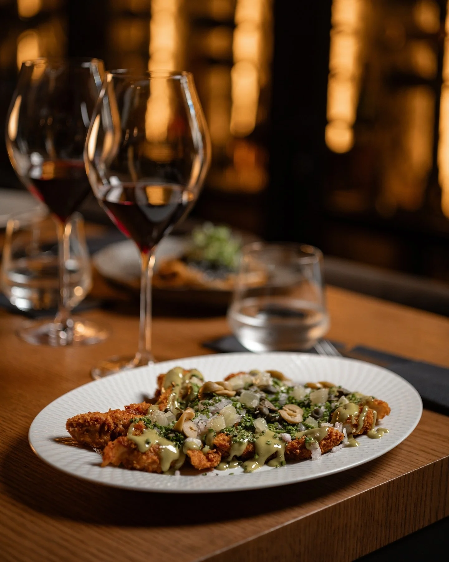 A plate of fried chicken topped with chopped onions, herbs, and drizzled with a green sauce, set on a wooden table with glasses of red wine and water, in a dimly lit restaurant.