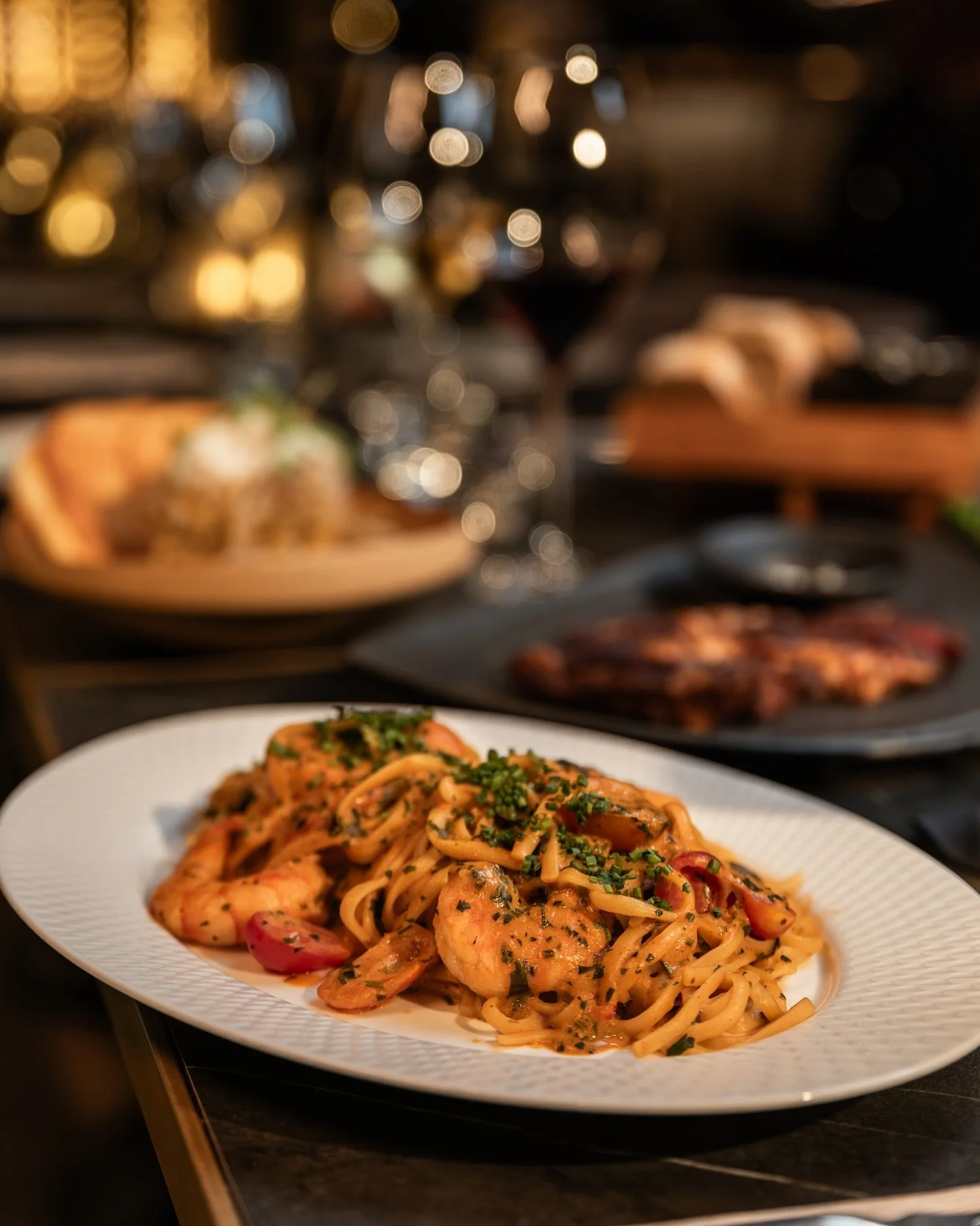 A plate of shrimp pasta garnished with chopped herbs, with wine glasses and other dishes blurred in the background.