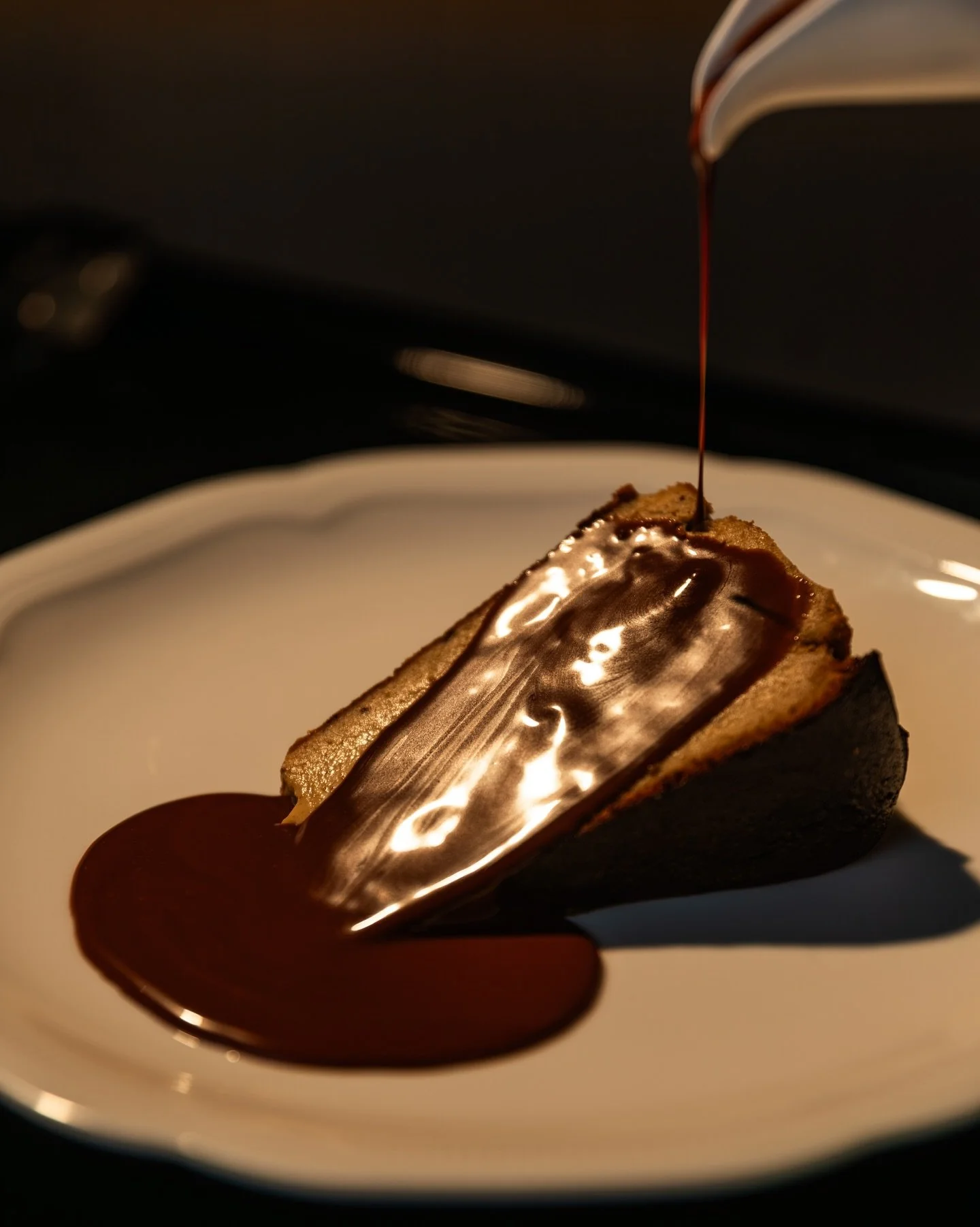 Slice of cheesecake with chocolate sauce and pouring chocolate over it on a white plate