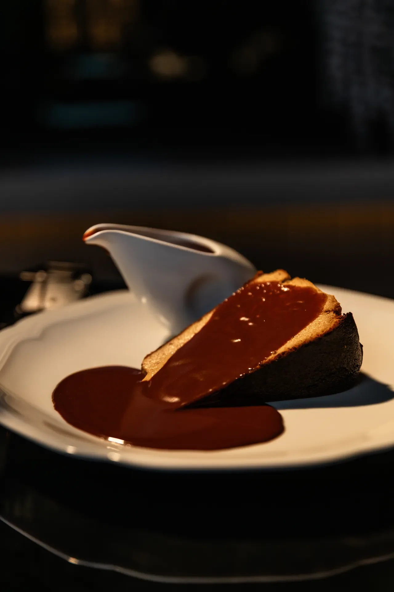 A dessert of a slice of cake with chocolate sauce on a white plate, next to a small white gravy boat.