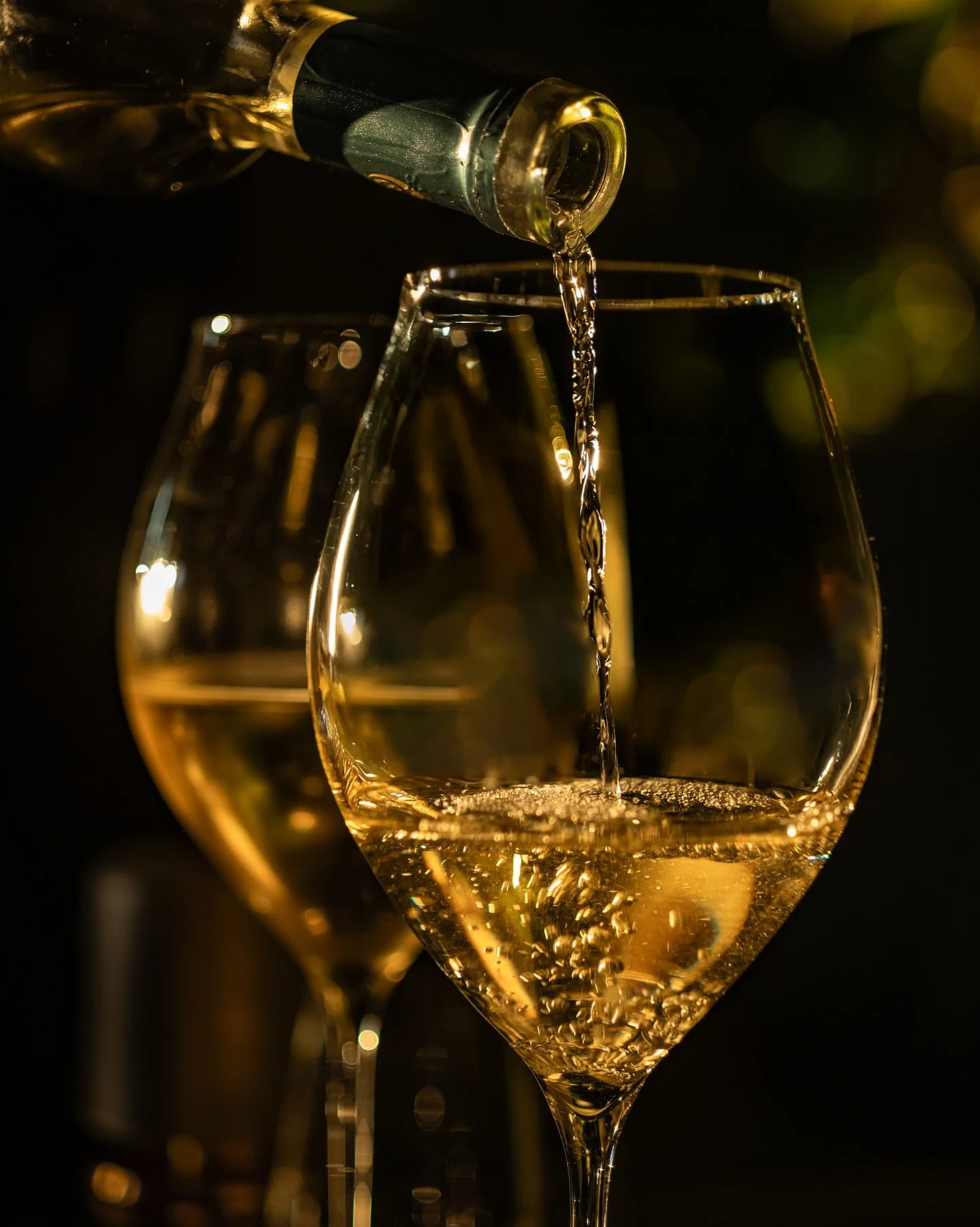 A close-up of white wine being poured from a bottle into a wine glass, with another glass in the background, illuminated by warm lighting.