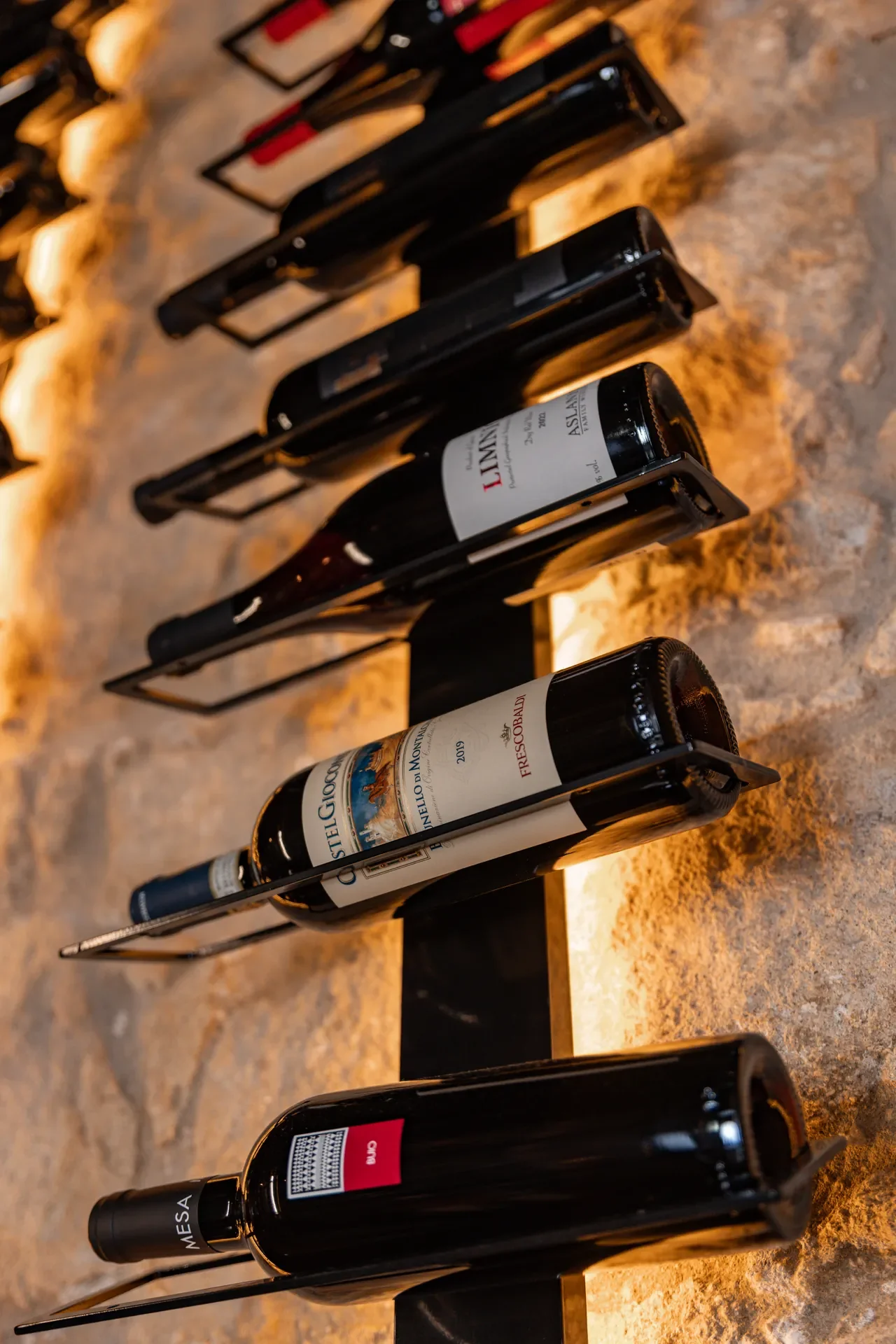 Multiple wine bottles displayed on a metal rack against a stone wall background.
