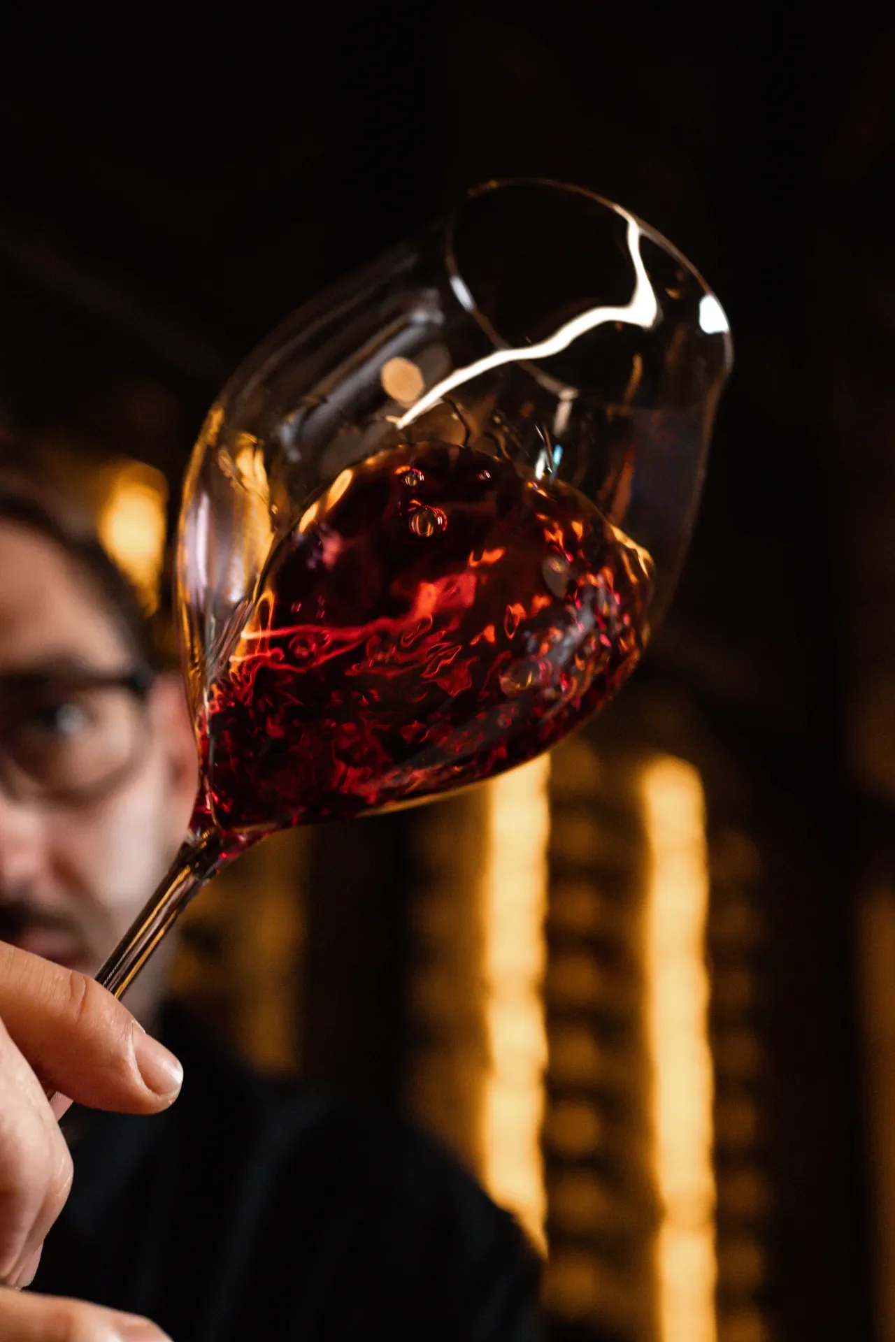 Person holding a glass of red wine with a dark background and blurred warm lights.