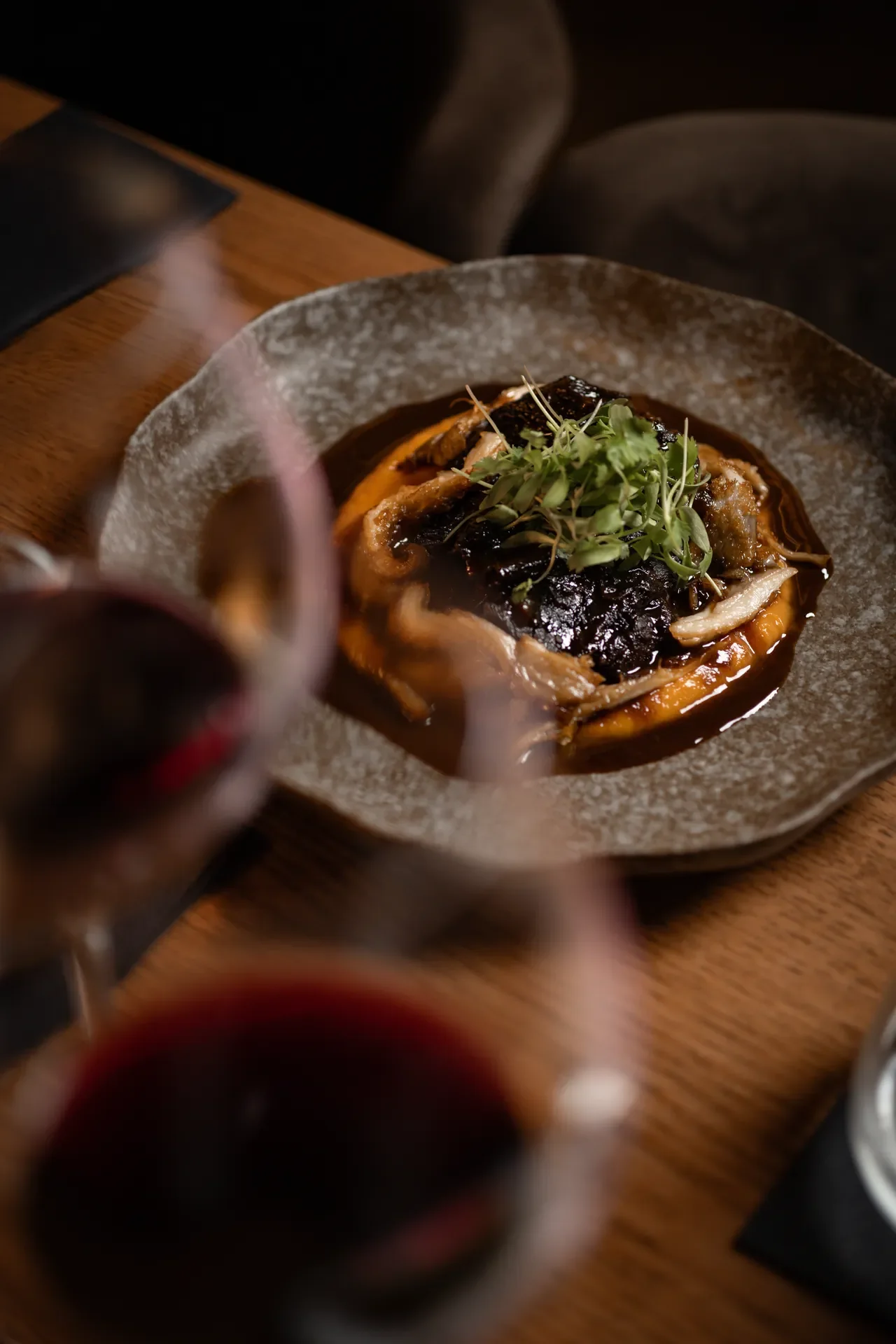 A gourmet dish with dark sauce, garnished with microgreens, served on a rustic ceramic plate, with glasses of red wine visible in the foreground