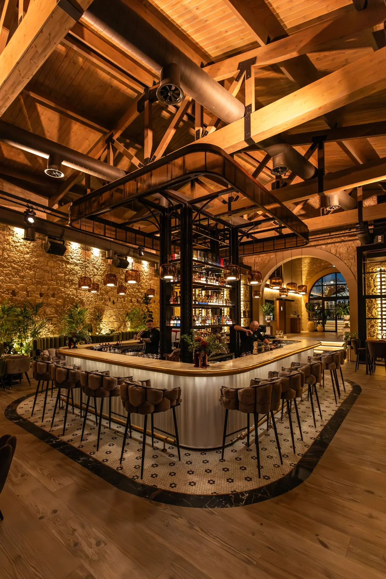 A stylish bar with a curved white counter, surrounded by bar stools, in a warm, industrial-style interior with exposed brick walls, wooden ceiling beams, and ambient lighting.