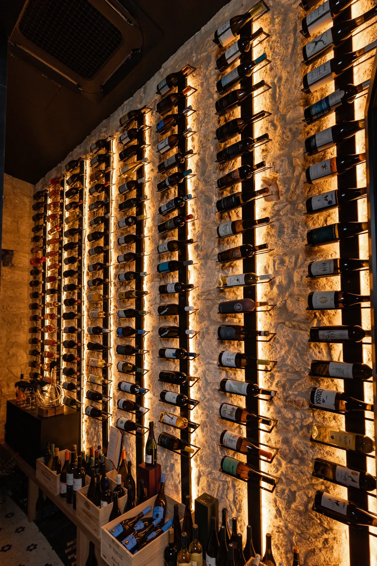 A wall displaying multiple rows of wine bottles illuminated with warm lighting at a wine shop or bar.