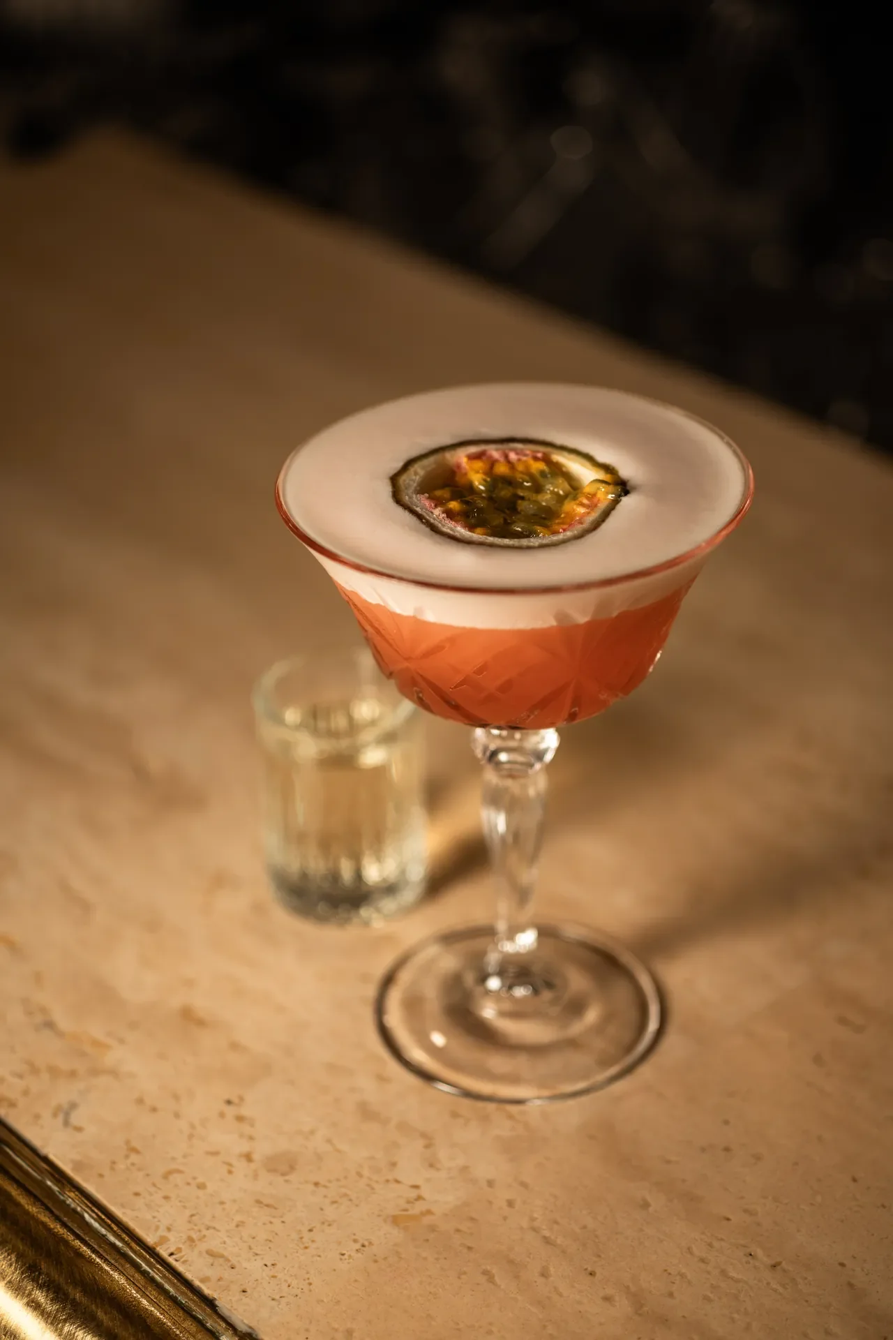 A cocktail in a vintage-style glass garnished with a passion fruit ring, on a marble bar surface.