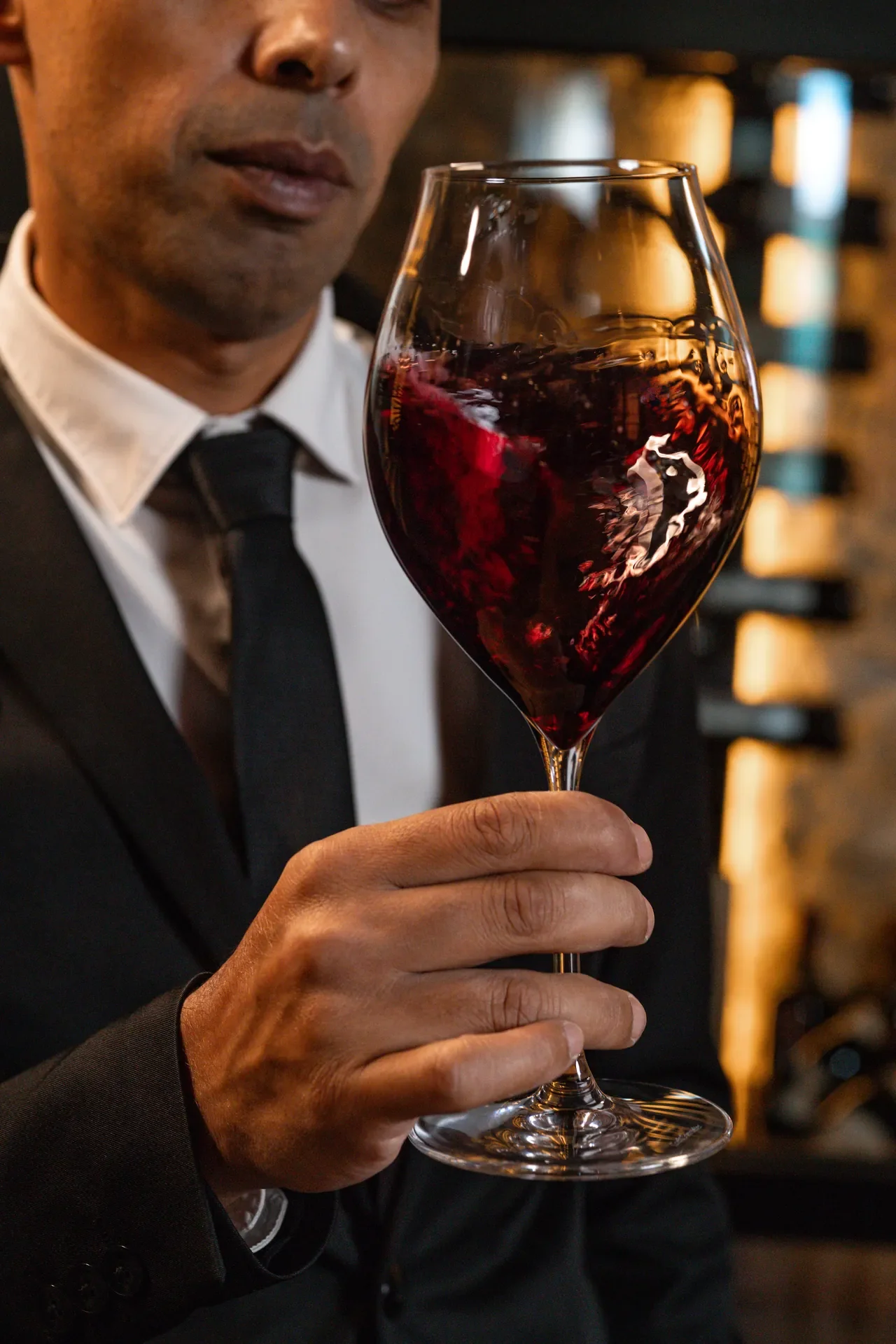 A man dressed in a black suit, white shirt, and black tie holding a glass of red wine.