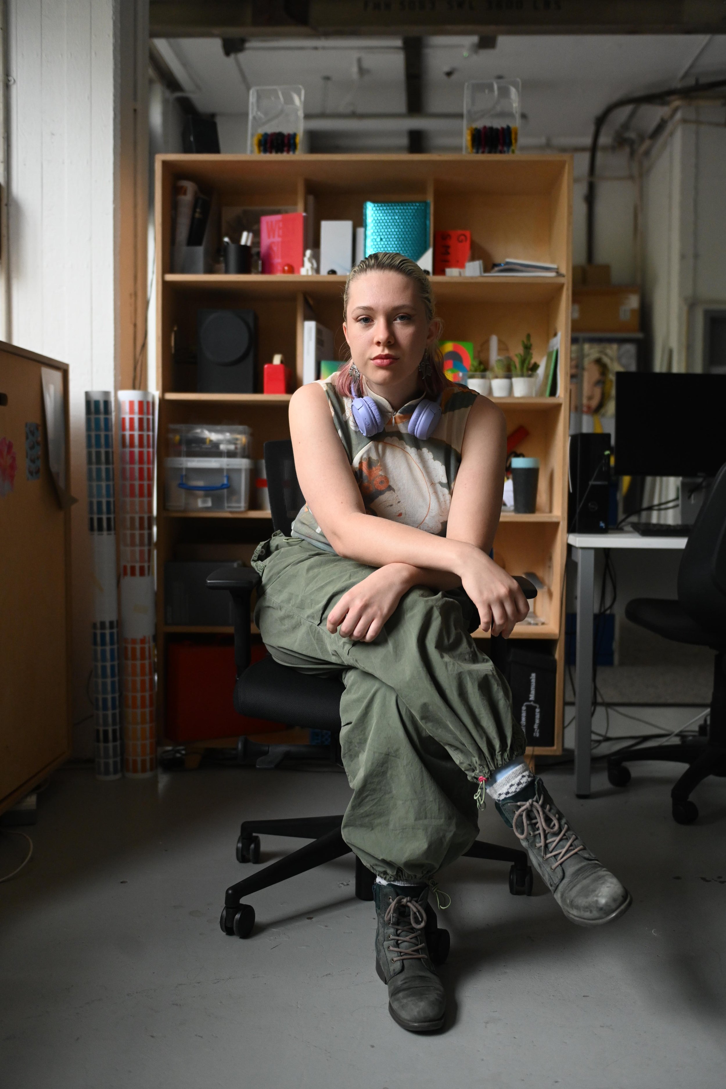 A young woman with pink hair sitting on an office chair in a room with shelves and office equipment, wearing a sleeveless shirt, green cargo pants, and hiking boots, with lavender headphones around her neck.