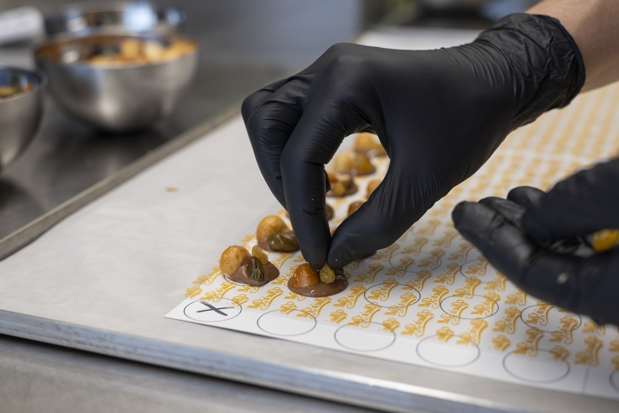 Eine Hand in schwarzen Handschuhen platziert kleine Lebensmittelstücke auf einen beschrifteten Untergrund in einer Küche.