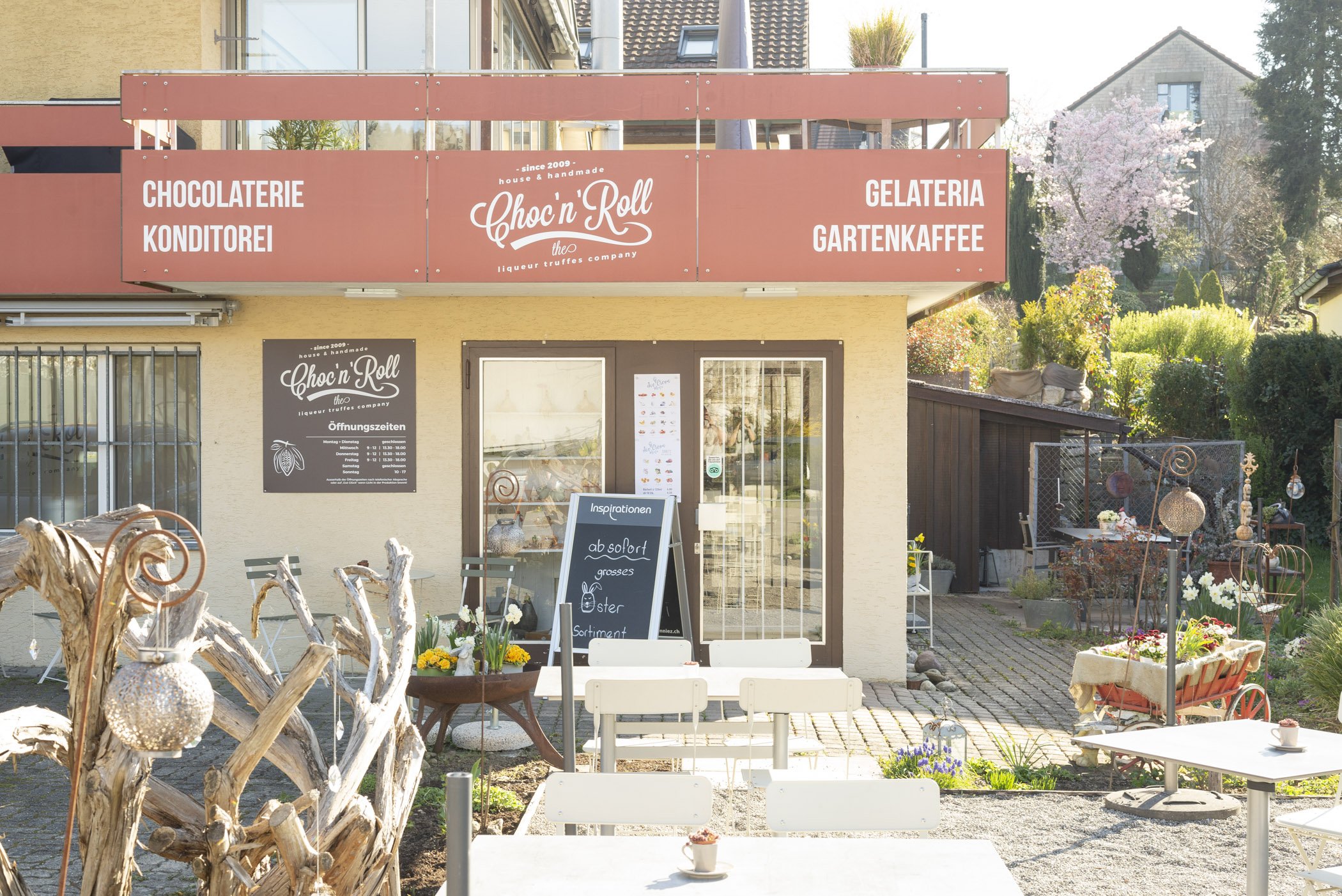 Außenansicht einer Chocolaterie und Konditorei mit Gartenkaffee, Sitzbereich im Freien, Dekorationen, Blumen und einem Schild mit Öffnungszeiten.