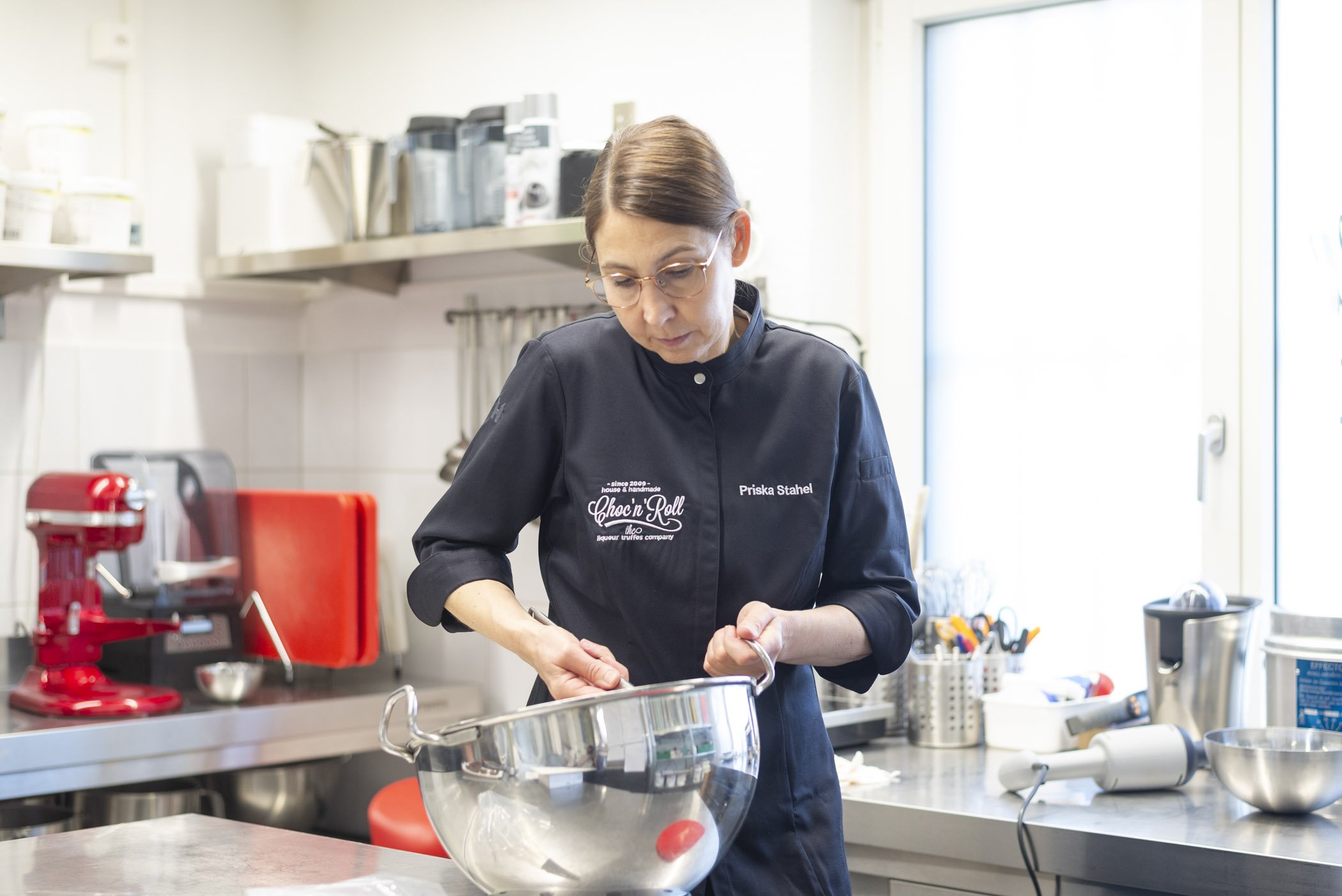 Frau in einer Küche, die in einer großen Metallrührschüssel arbeitet. Sie trägt eine schwarze Küchenjacke mit einem Namensschild. Im Hintergrund befindet sich ein roter Mixer und Küchenutensilien.