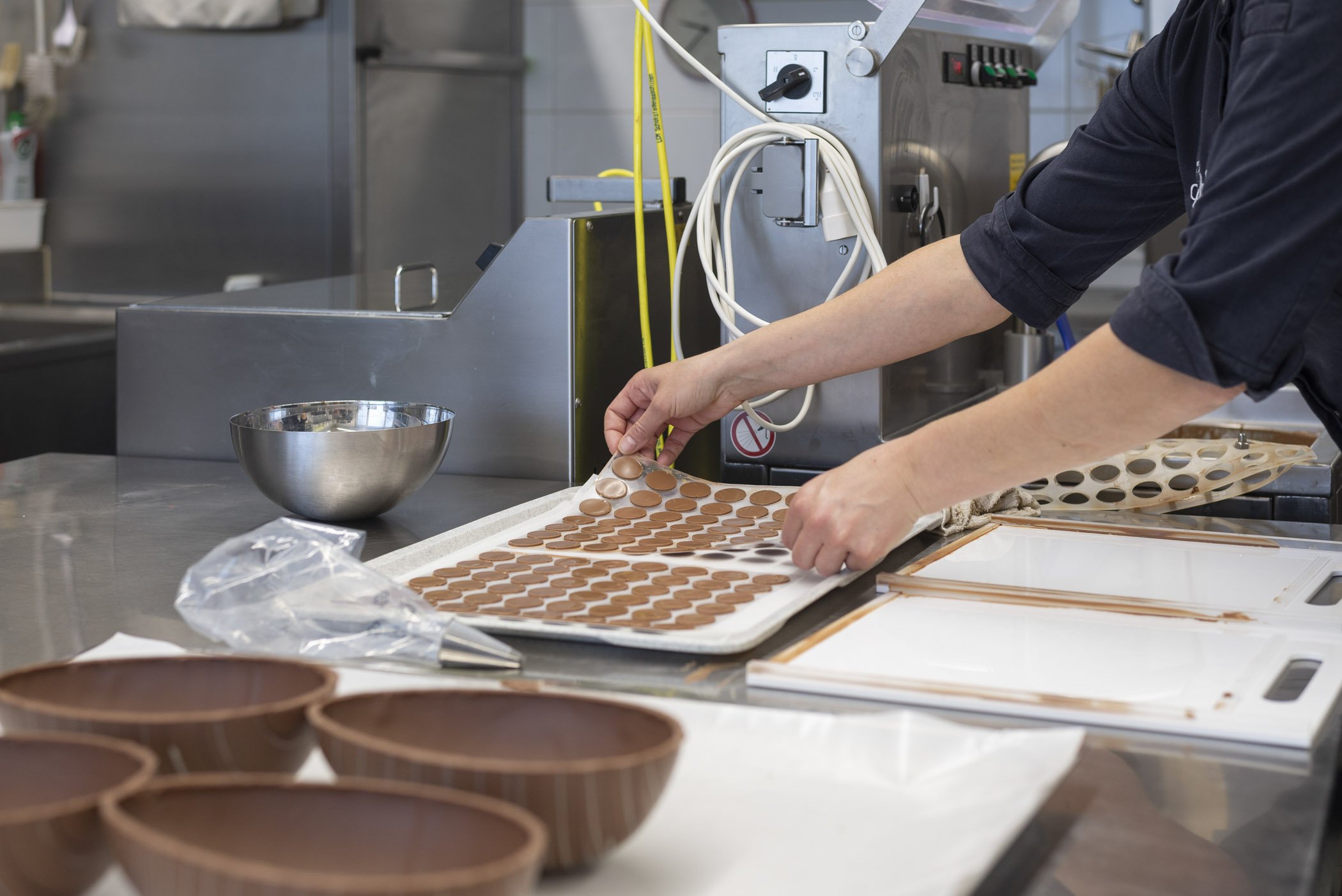 Person in einer Konditorei oder Schokoladenwerkstatt, die eine Form mit kleinen runden Schokoladenstücken herstellt. Auf dem Tisch sind auch Halbschalen aus Schokolade und ein Rührgerät zu sehen.
