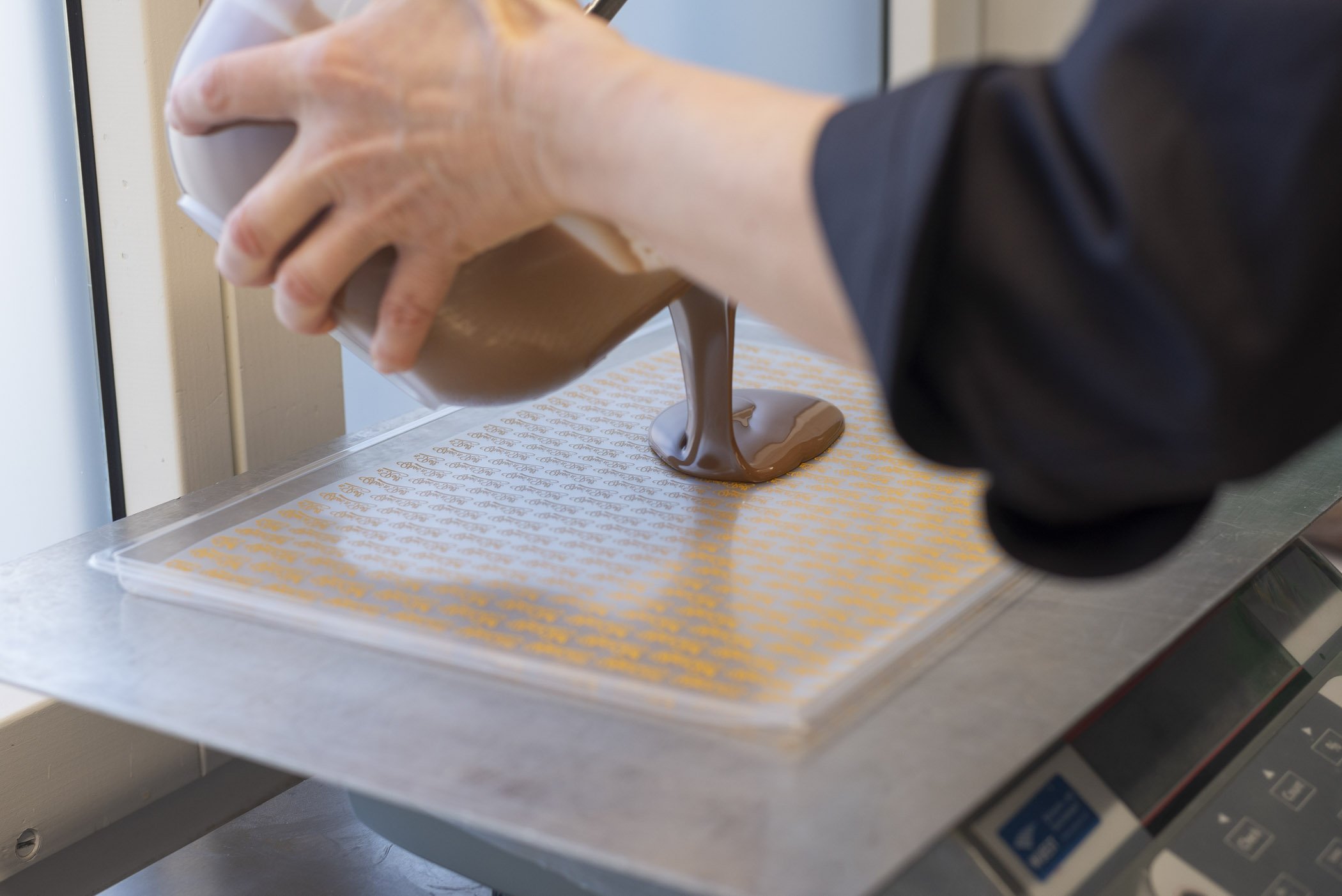 Person gießt flüssige Schokolade auf ein Tablett mit Schablone in einer Küche.