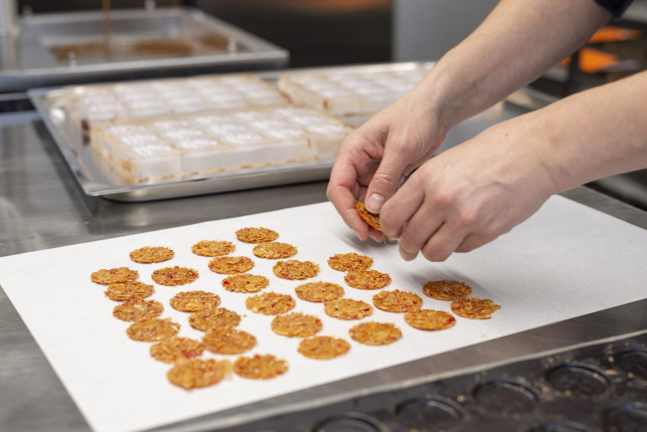 Person arrangiert kleine, runde Keksplätzchen auf Backpapier in einer Bäckereiumgebung.