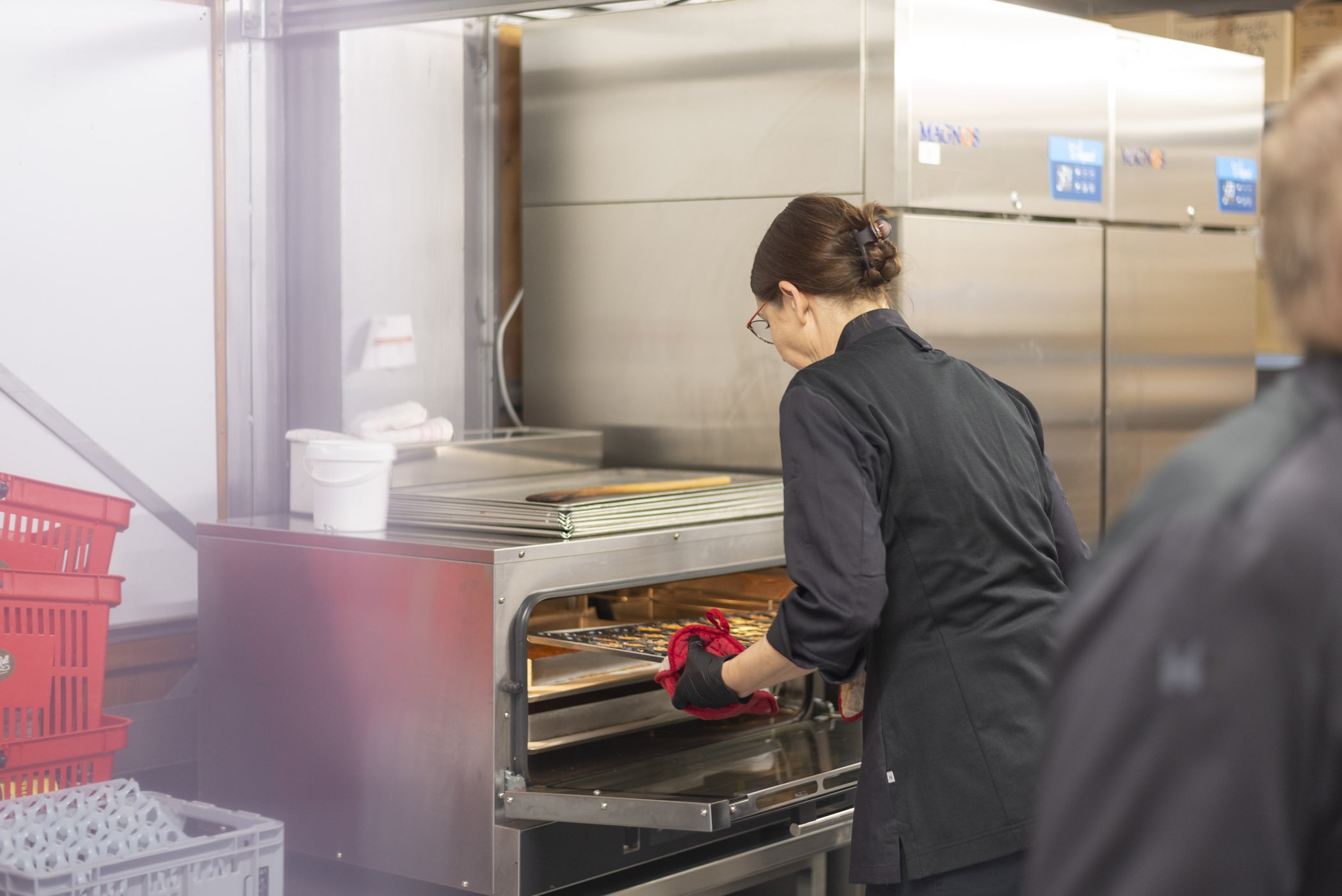 Eine Person in schwarzer Arbeitskleidung öffnet einen Ofen und nimmt ein Backblech heraus. In der Umgebung sieht man industrielle Küchenutensilien und Geräte.