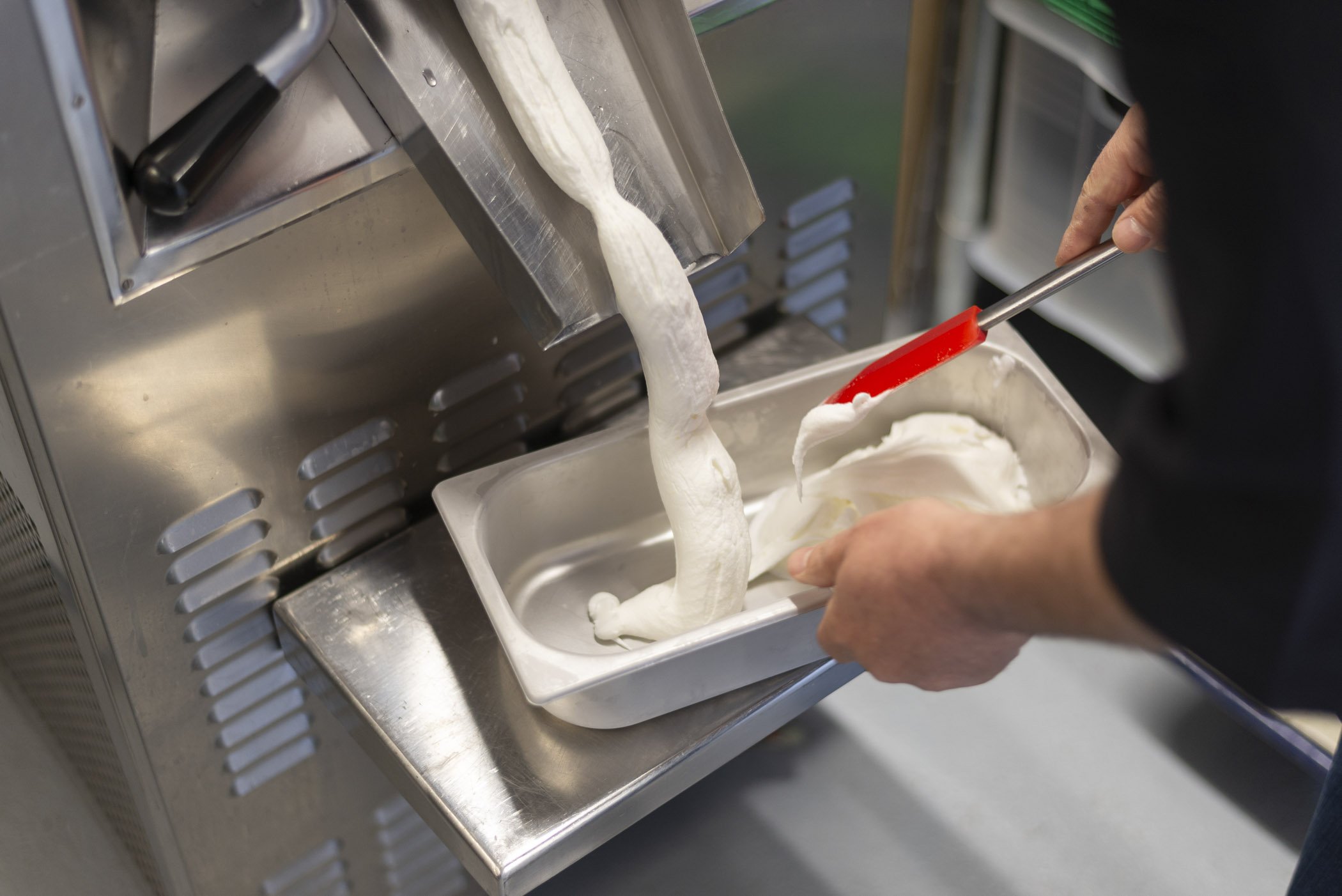 Zubereitung von Eiscreme mit einer Eismaschine, wobei eine Person mit einem Spatel Eis in einen Metallbehälter füllt.